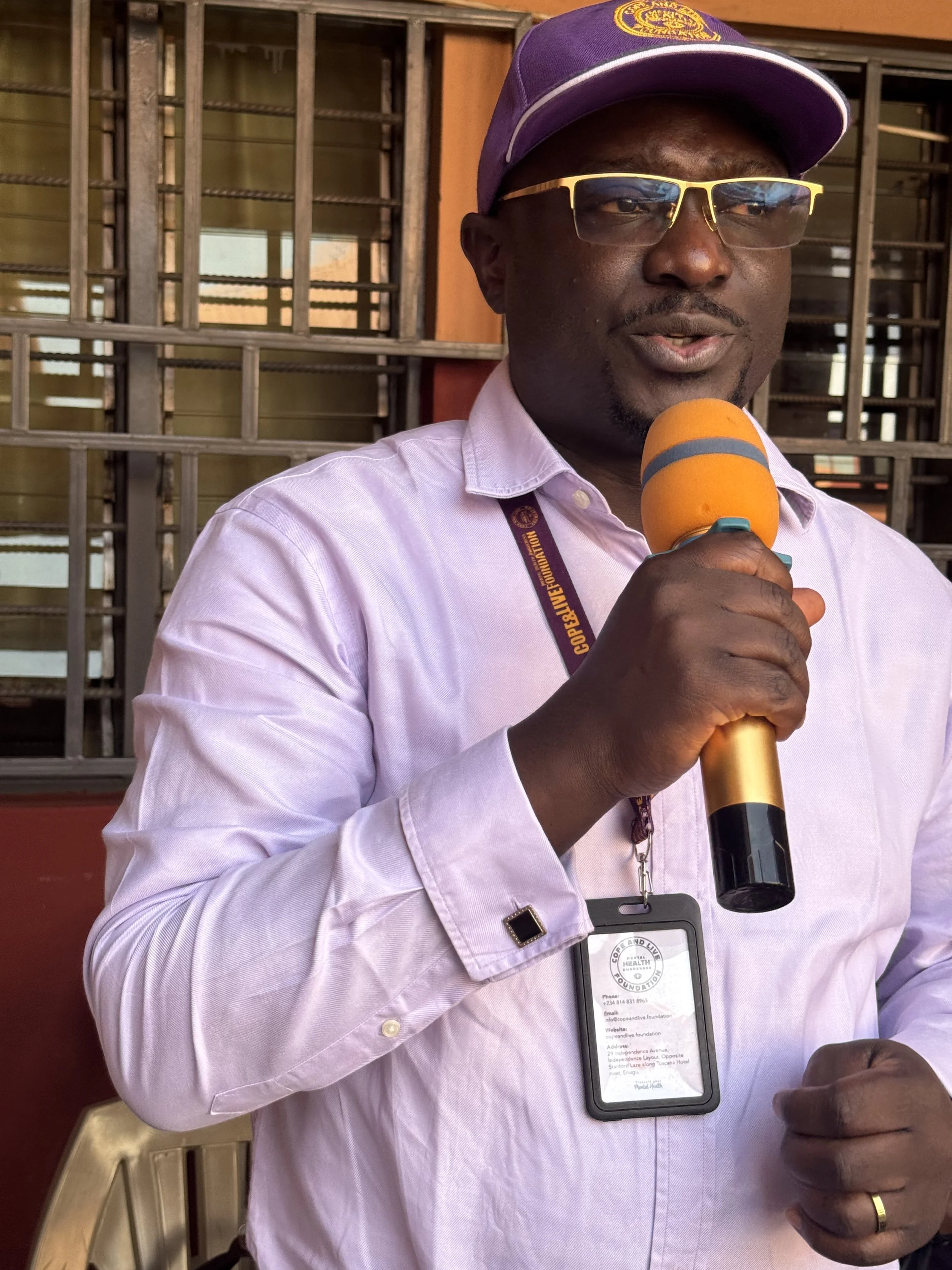 A man wearing a purple cap, glasses, and a light pink shirt is speaking into an orange microphone. He has a badge hanging around his neck.