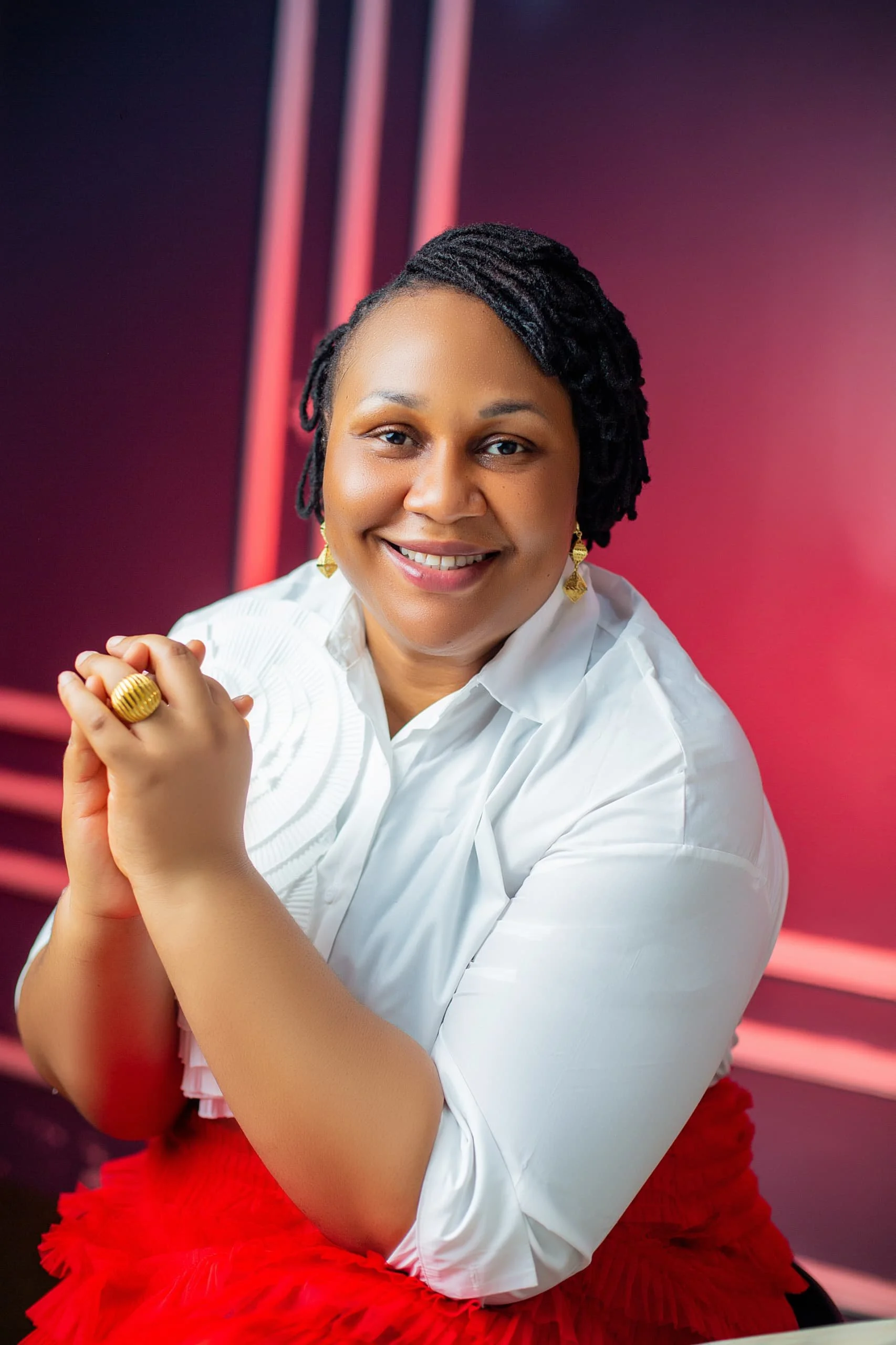 A smiling woman with short black twisted hair, wearing a white blouse, gold jewelry, and a red skirt, is posing with her hands clasped together against a pink and purple background with geometric lines.