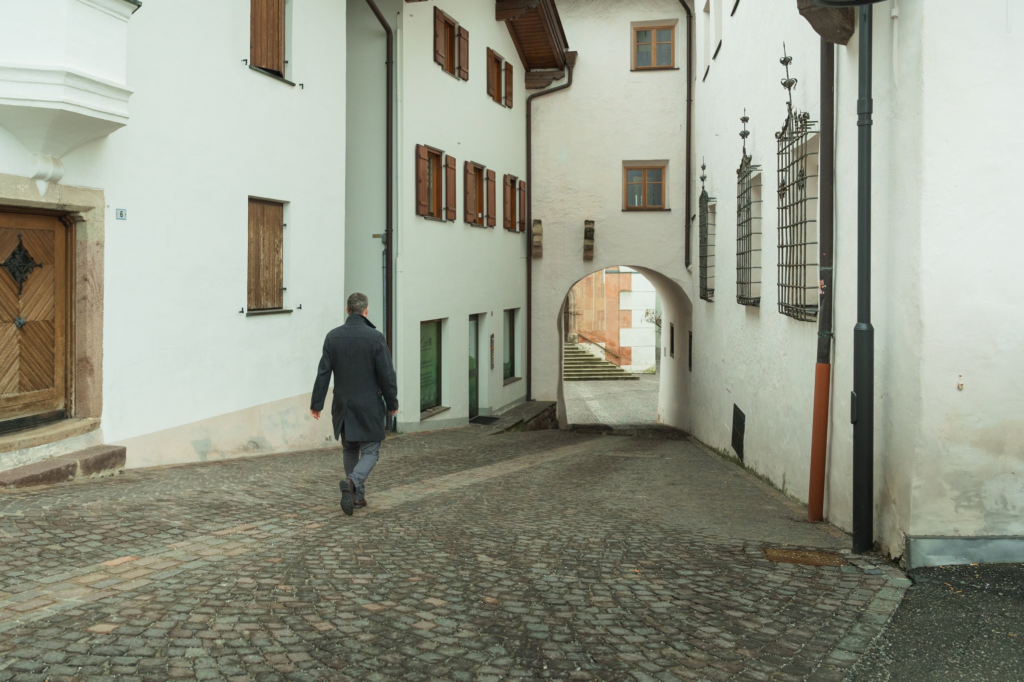 Rudi Sampr geht in einer engen, kopfsteingepflasterten Gasse einer Altstadt, umgeben von weißen Gebäuden mit Holztüren und Fenstern. Er geht zu seinem Büro in Kastelruth, Südtirol.