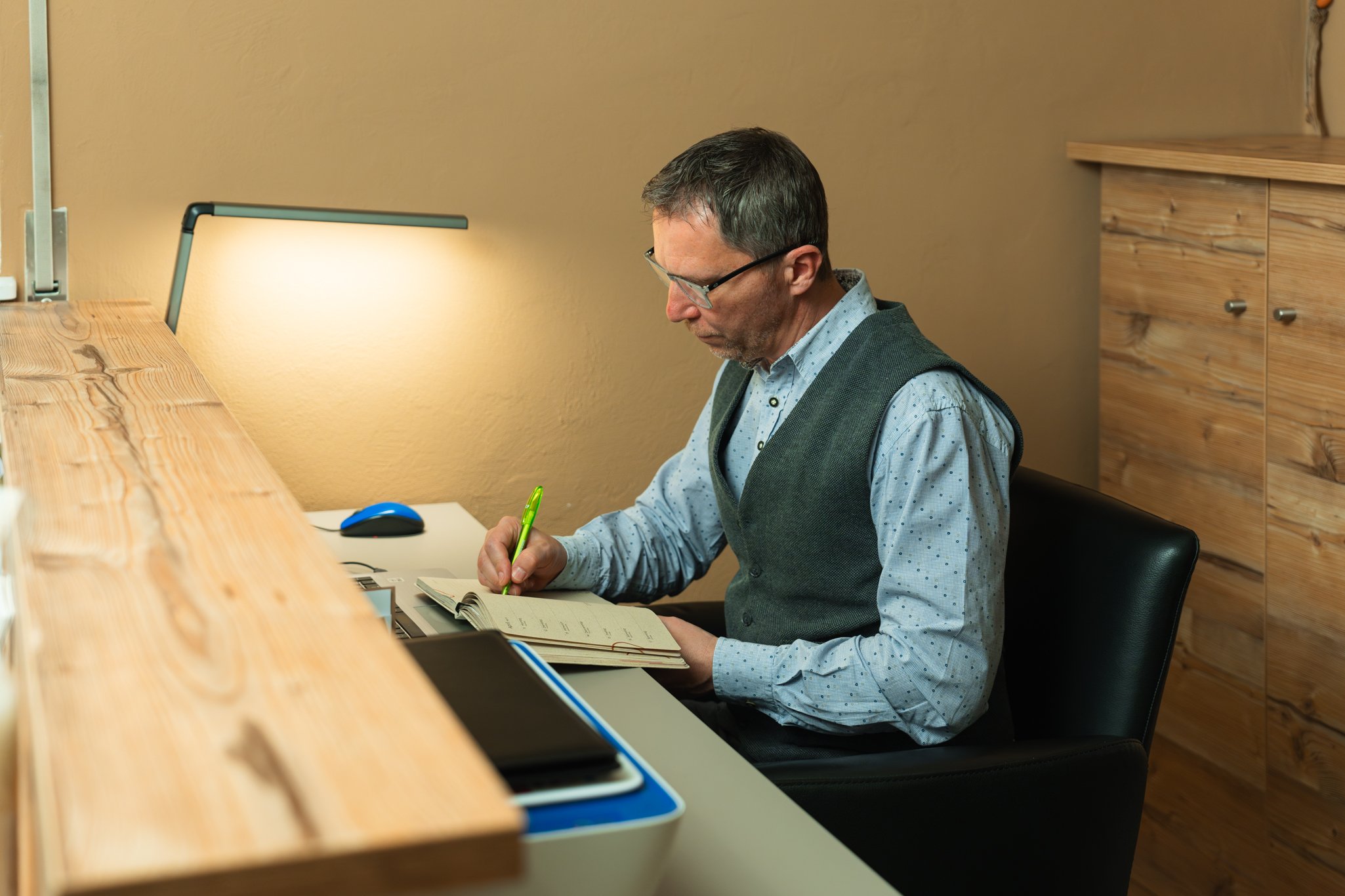 Rudi Sampt sitzt an einem Schreibtisch und schreibt in ein Notizbuch, umgeben von Holzverkleidung und einer Tischlampe. Er hält ein online Meeting mit einem Klienten.