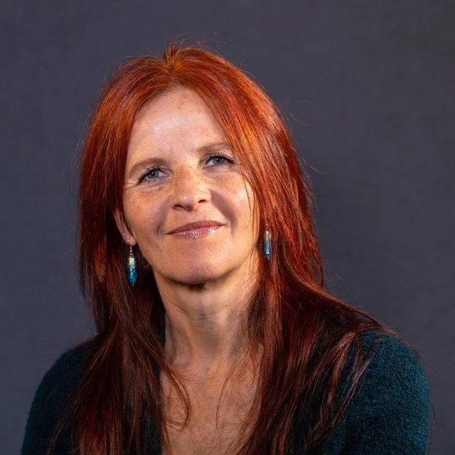 Porträt einer Frau mit roten Haaren, blaue Ohrringe, grauem Hintergrund, lächelnd.