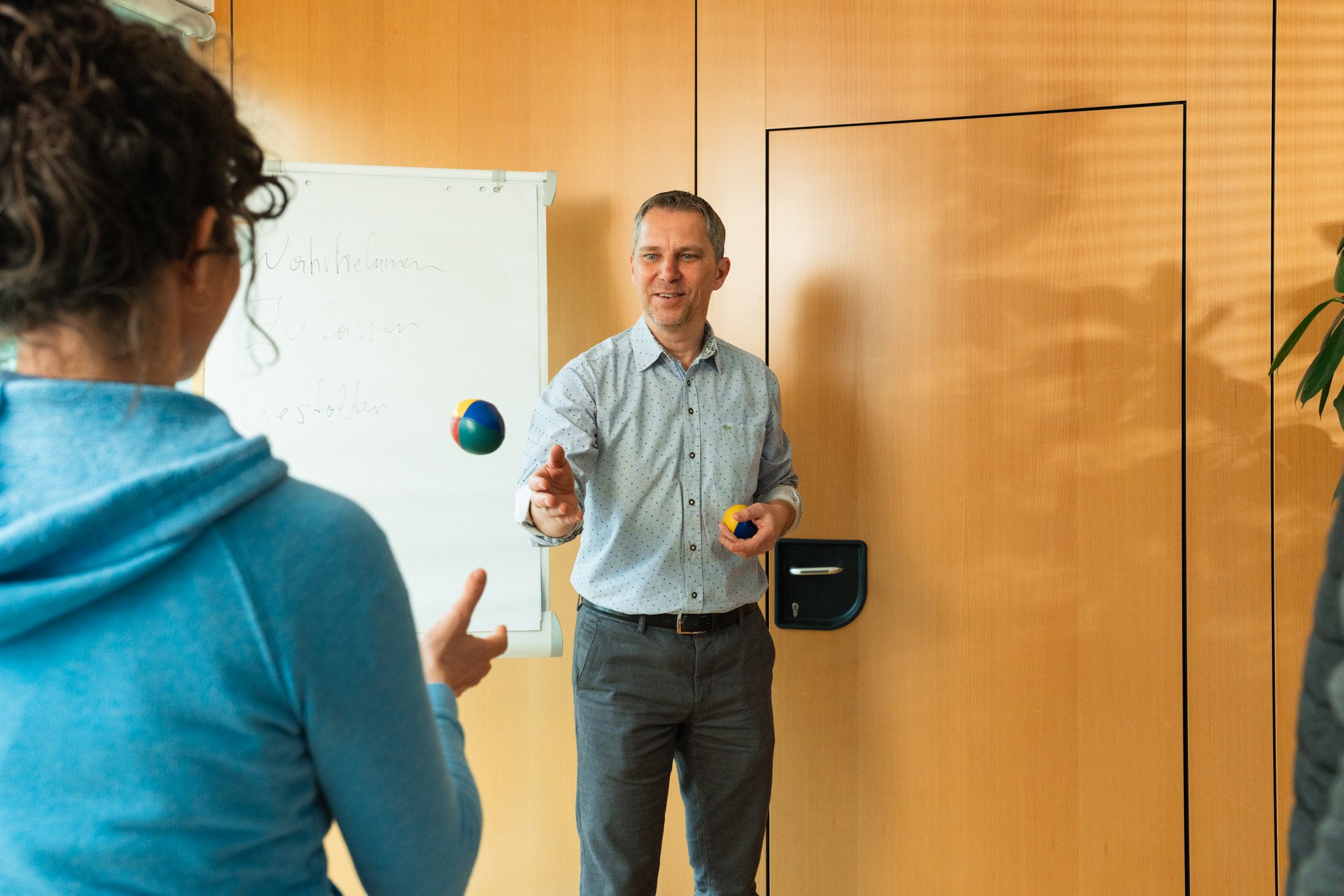 Zwei Personen jonglieren Bälle im Raum mit einer Flipchart im Hintergrund. Es handelt sich um ein Konzentrationsspiel.