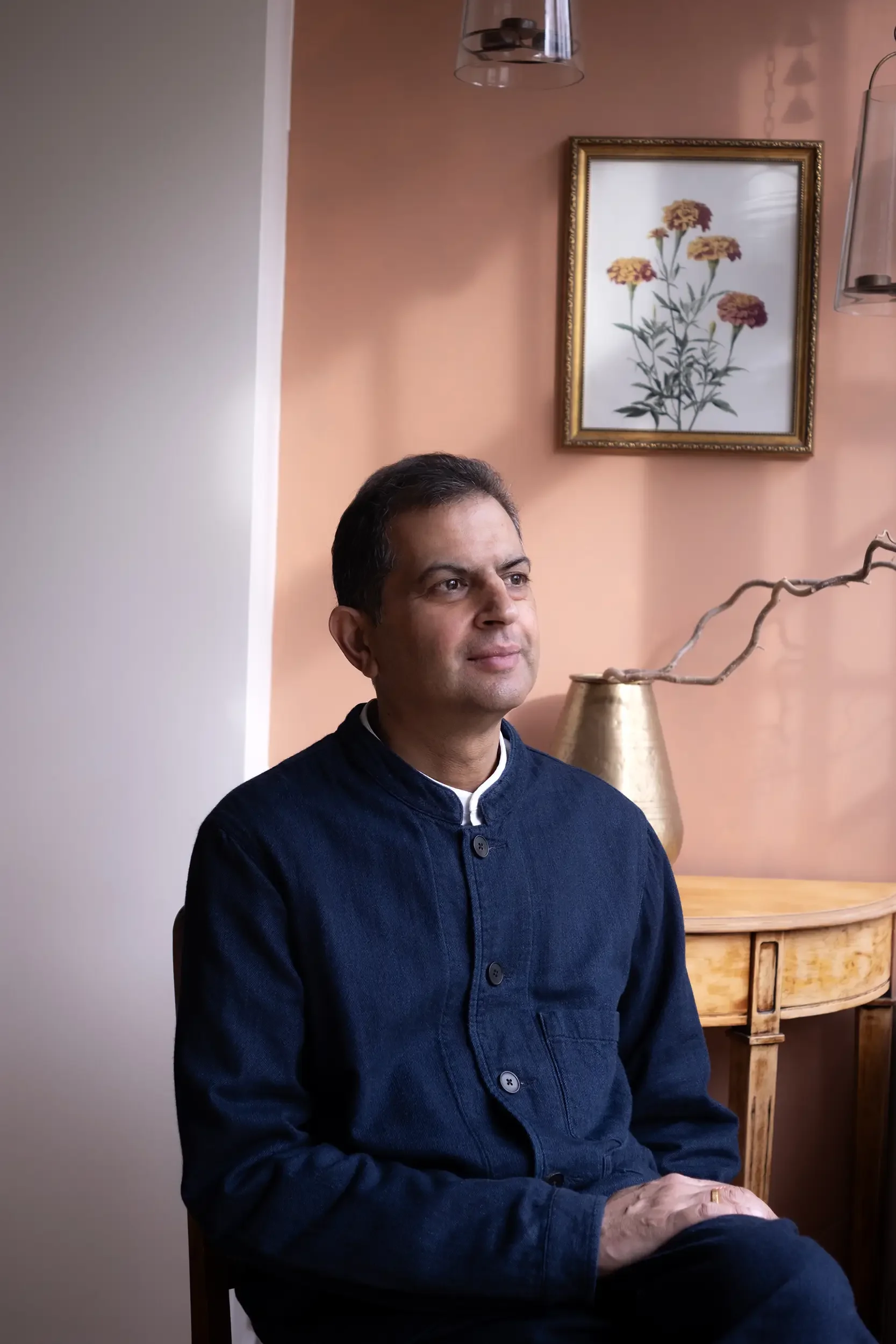 A man sitting indoors in front of a pink painted wall with a framed flower picture and a decorative plant on a wooden table.