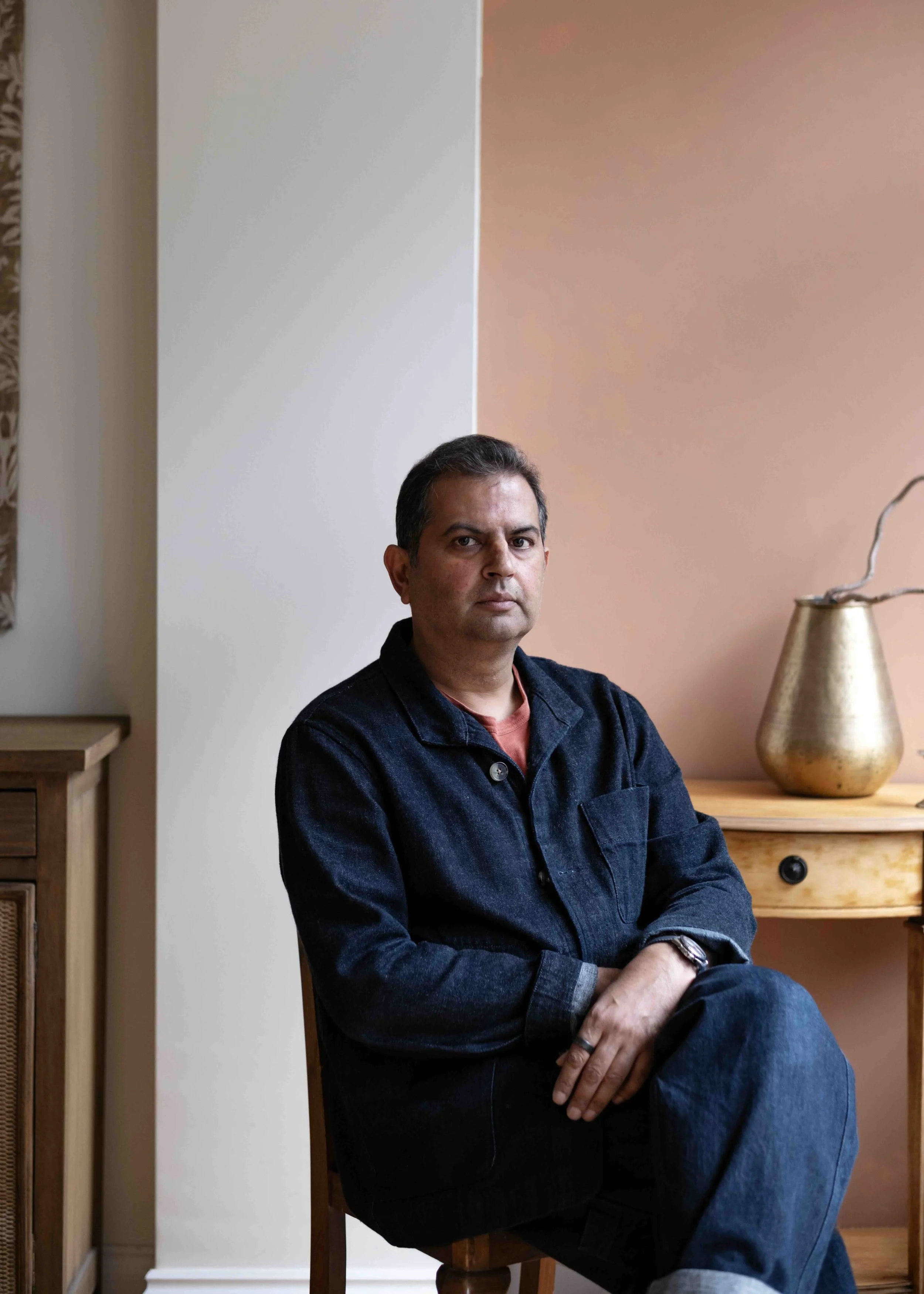 A man sitting on a wooden chair in a room with peach and white walls, wearing a black denim jacket and dark jeans, with a gold-colored vase on a wooden side table behind him.