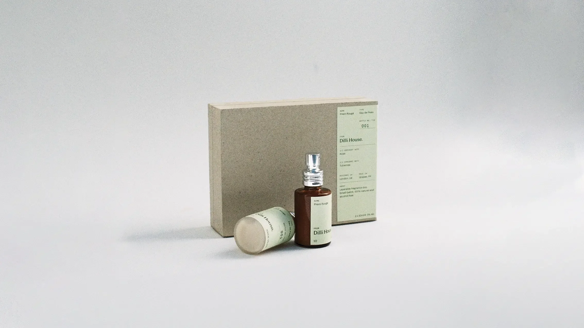 A small amber glass spray bottle with a silver cap, laying next to a round lid, both in front of a beige box with green labeling, on a plain white background.