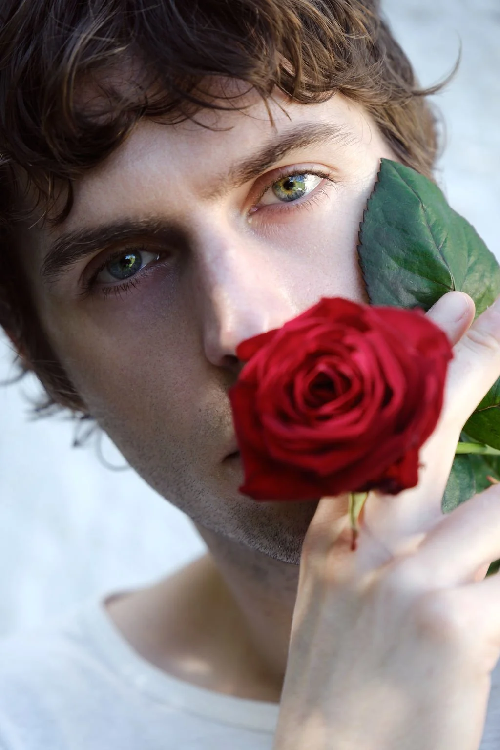 A man holding a red rose over his face, eyes meeting the camera