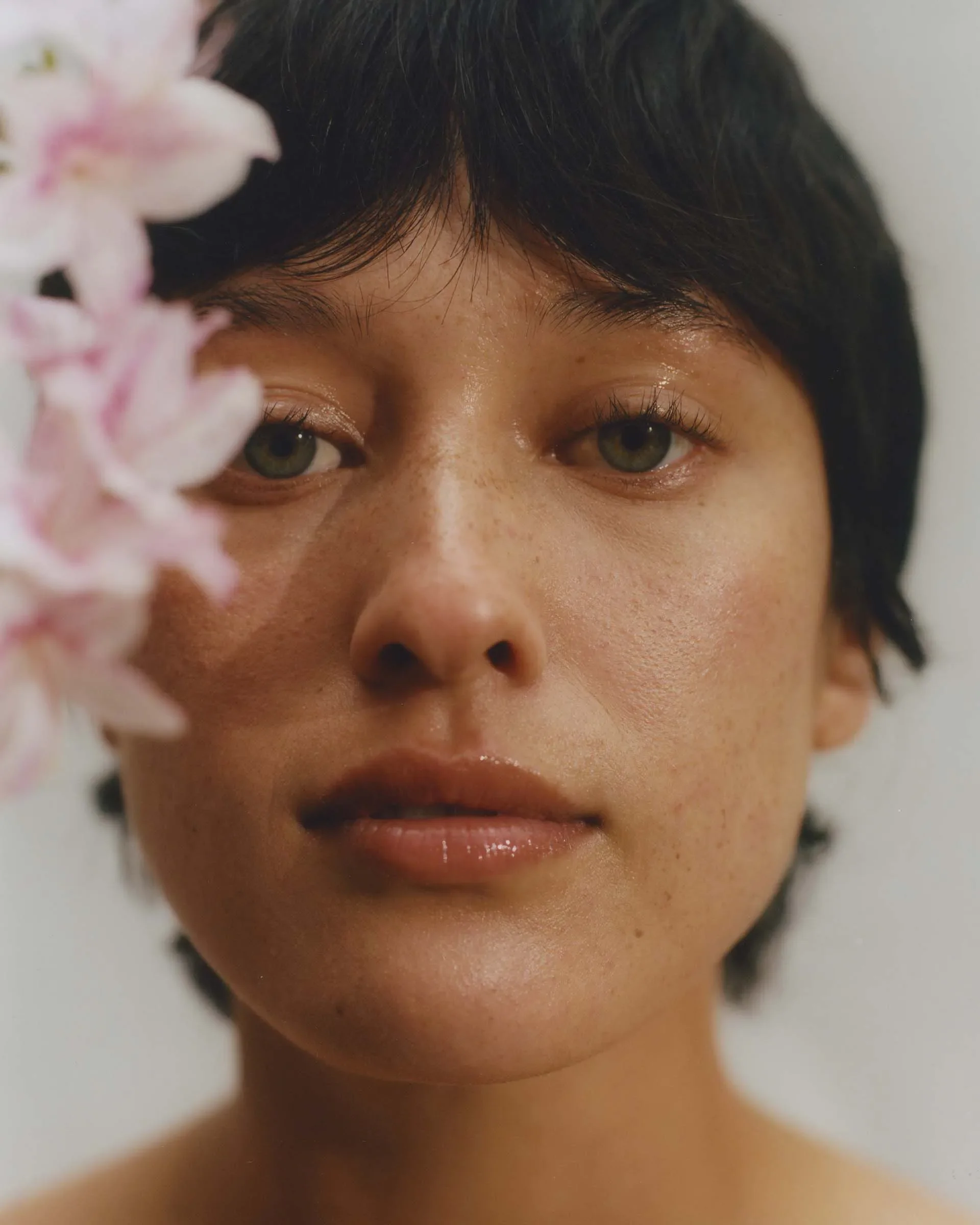 Close-up portrait of a person with short dark hair and light skin, partially obscured by pink flowers.