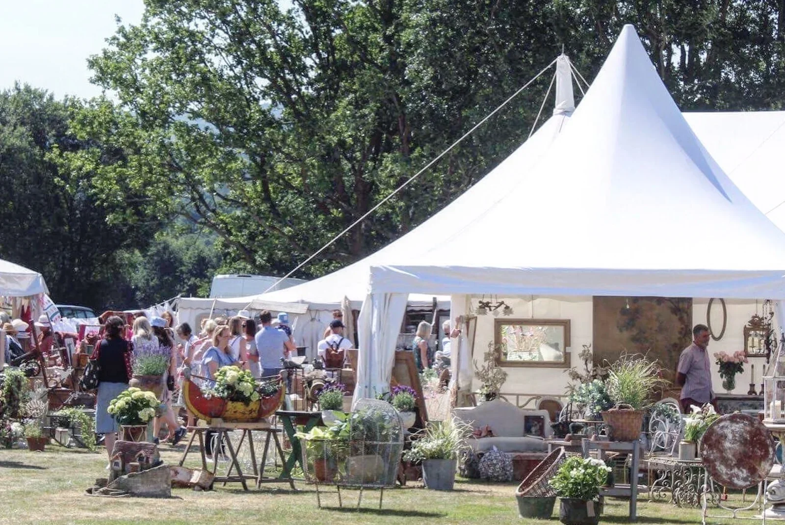 People browsing and shopping at an outdoor market with white tents, displaying vintage and antique items, surrounded by trees.