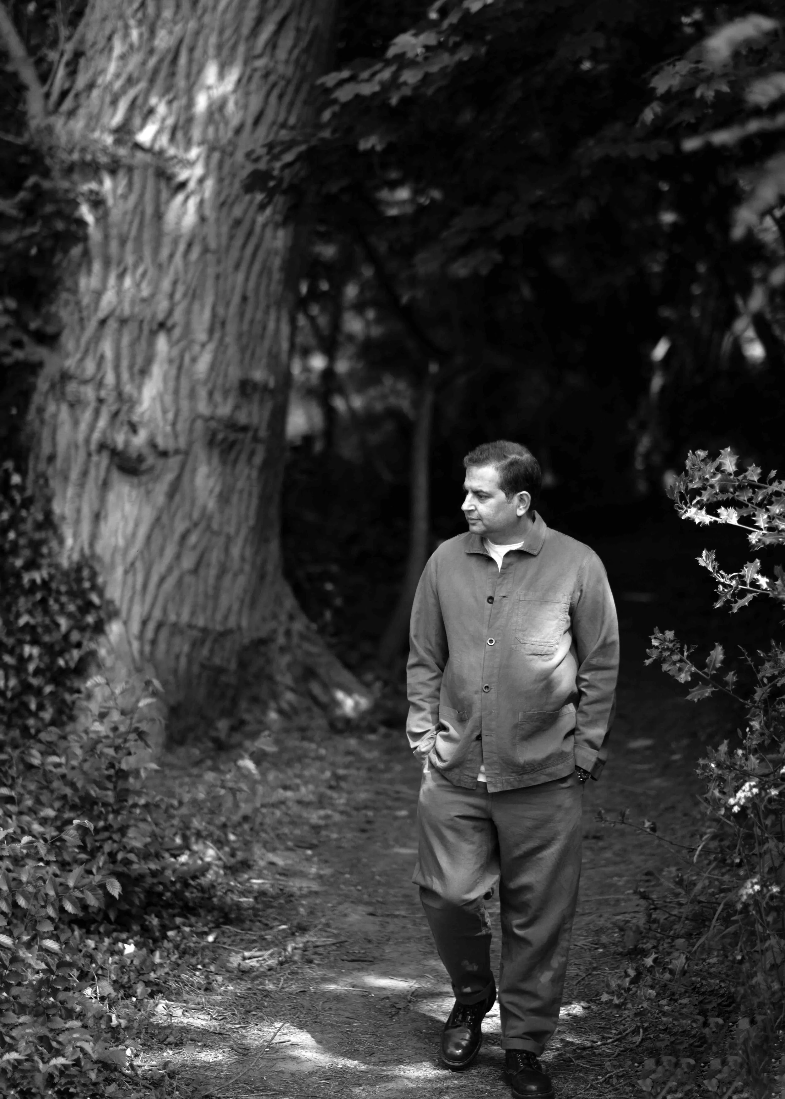 A man walking on a dirt path surrounded by trees and bushes in black and white.