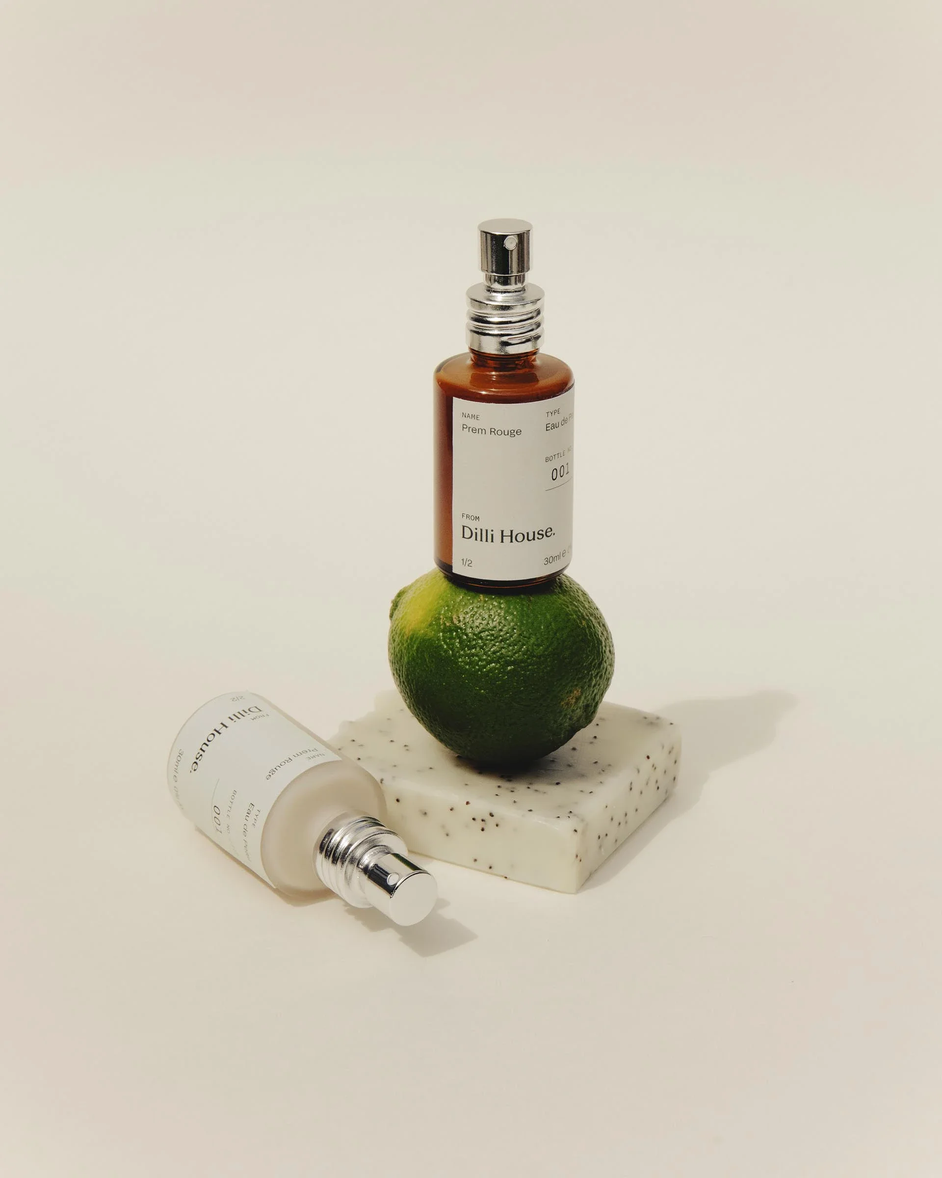 Two skincare bottles labeled 'Dill House' resting on an artificial lime, which is placed on a white speckled square stone, with one bottle upright and the other on its side.