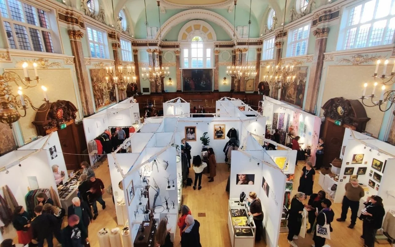 The main hall at Chelsea Old Town Hall during Handmade Chelsea, seen from the gallery: white exhibitor stands in rows on a wooden floor, visitors moving between them, chandeliers lit, and the hall's green ceiling and paintings above.
