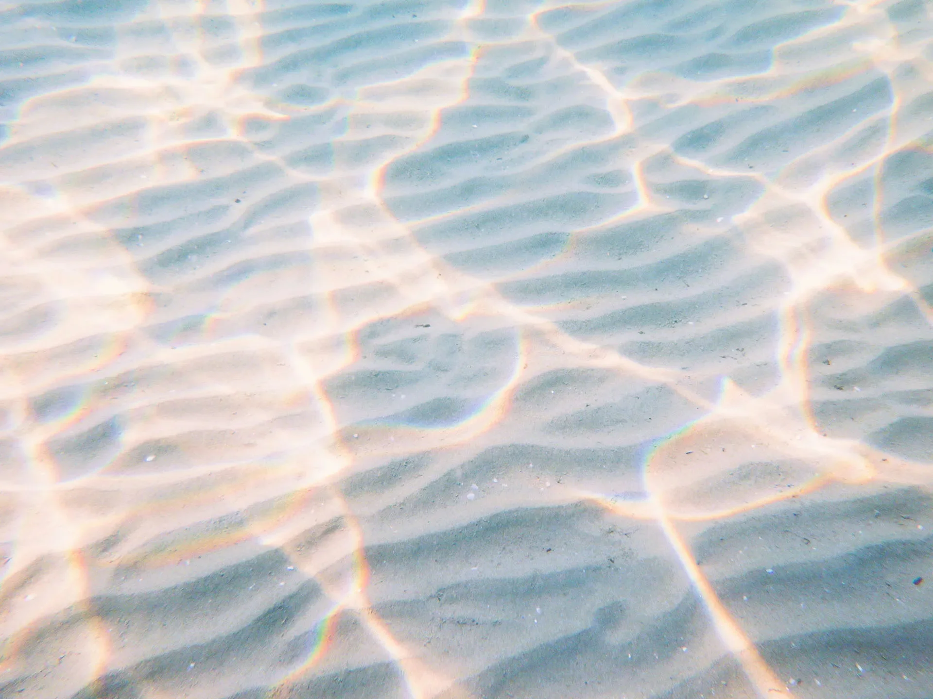 Shallow water at the beach with sunlight creating rainbow-colored patterns on the sandy bottom.