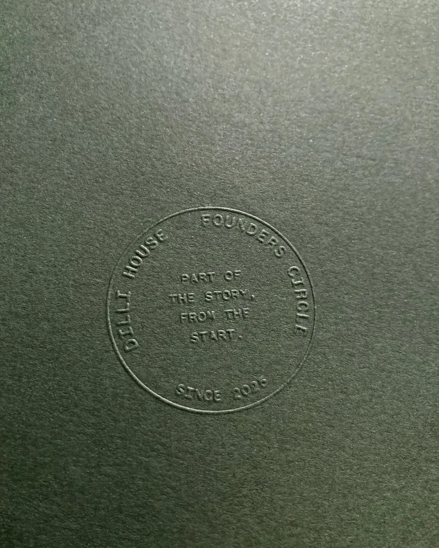Embossed circle on dark textured paper with the words, ' DILLI HOUSE FOUNDERS CIRCLE. PART OF THE STORY.  FROM THE START.  SINCE 2025.'