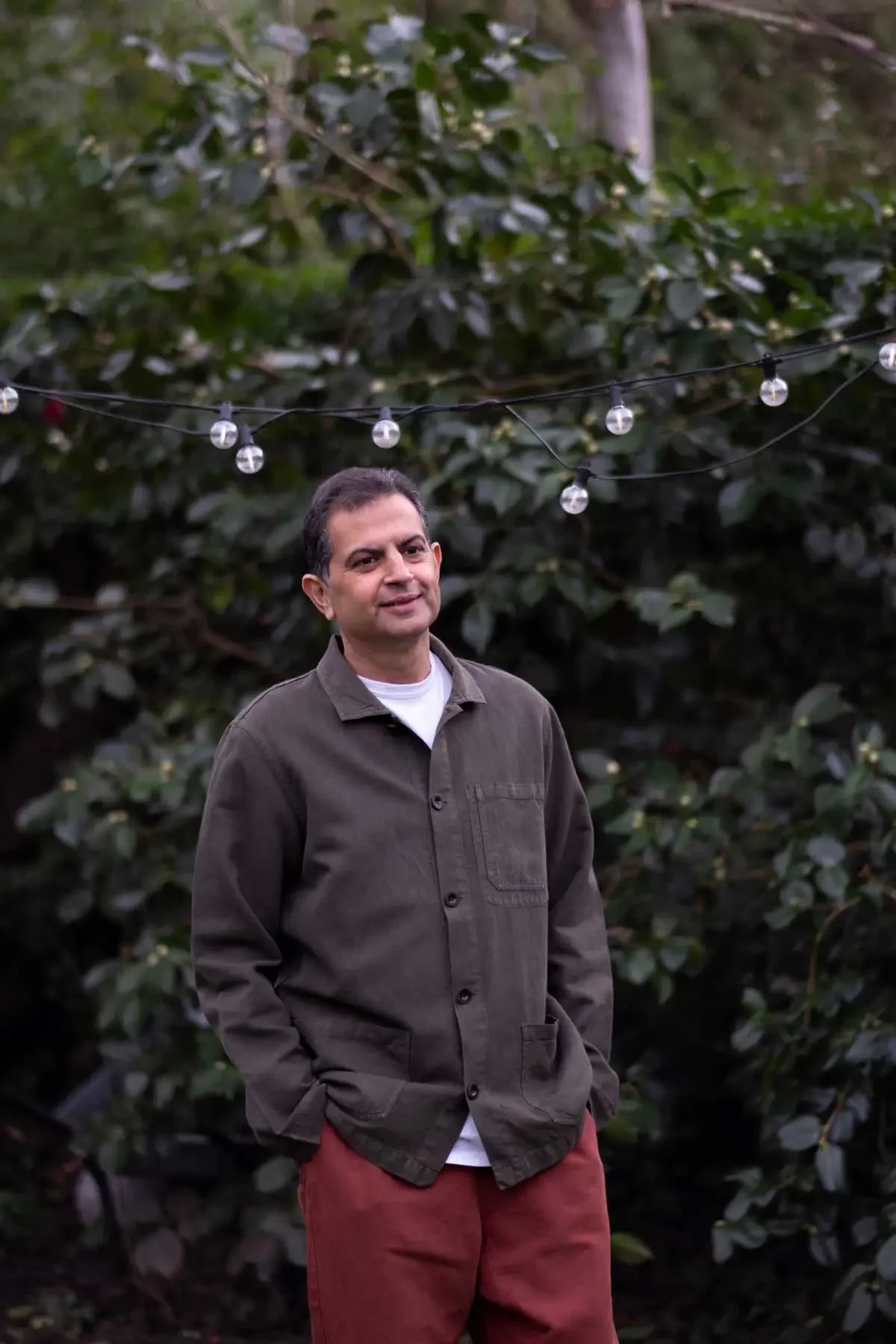 Man standing outdoors in front of dense green foliage, wearing a brown jacket and red pants, with string lights hanging overhead.