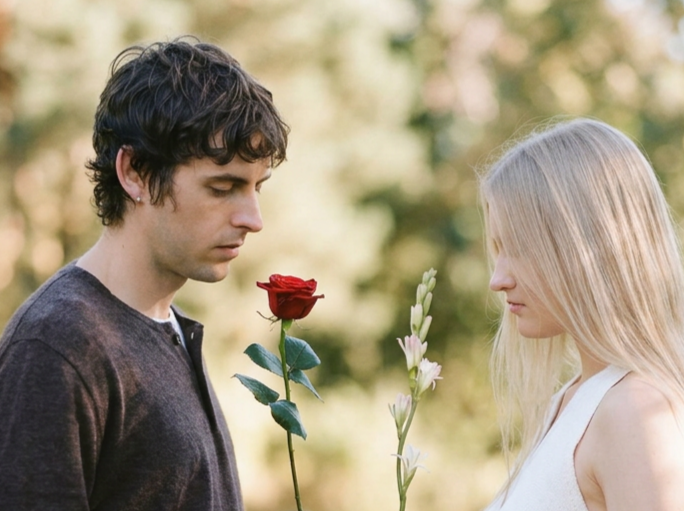 Man holding a red rose and woman holding a stem of tuberose, both looking down, outdoors in soft light