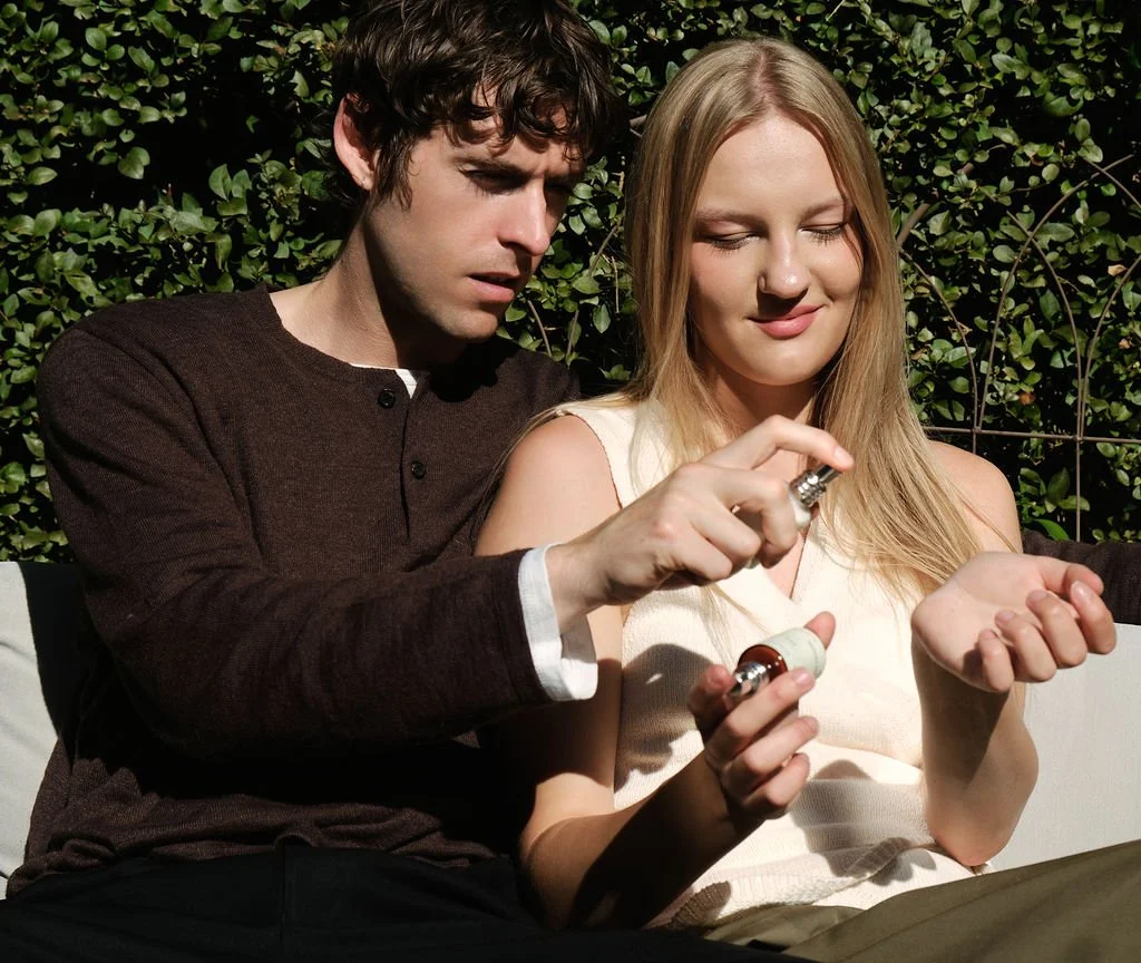 Two people layering Prem Rouge, one spraying the fragrance onto the other’s wrist in a garden