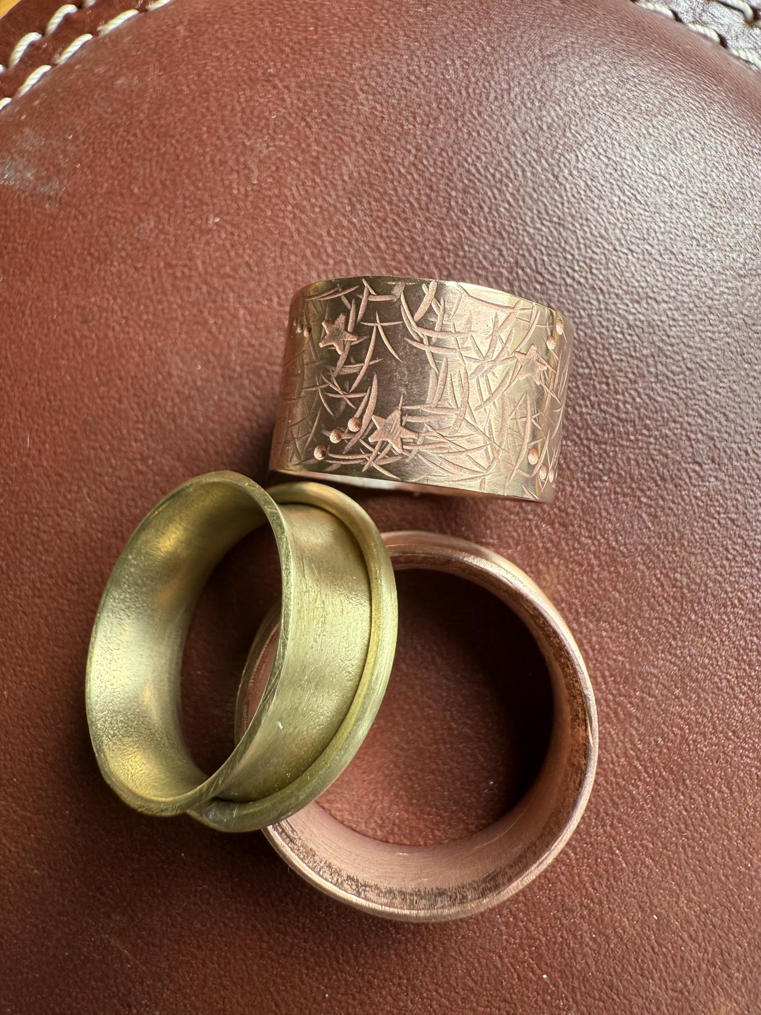 Three rings of different sizes and colors are placed on a reddish-brown textured surface. One ring is gold with a smooth surface, another is a pinkish bronze with a textured surface, and the third is a wide bronze or copper ring with engraved star and leaf patterns.