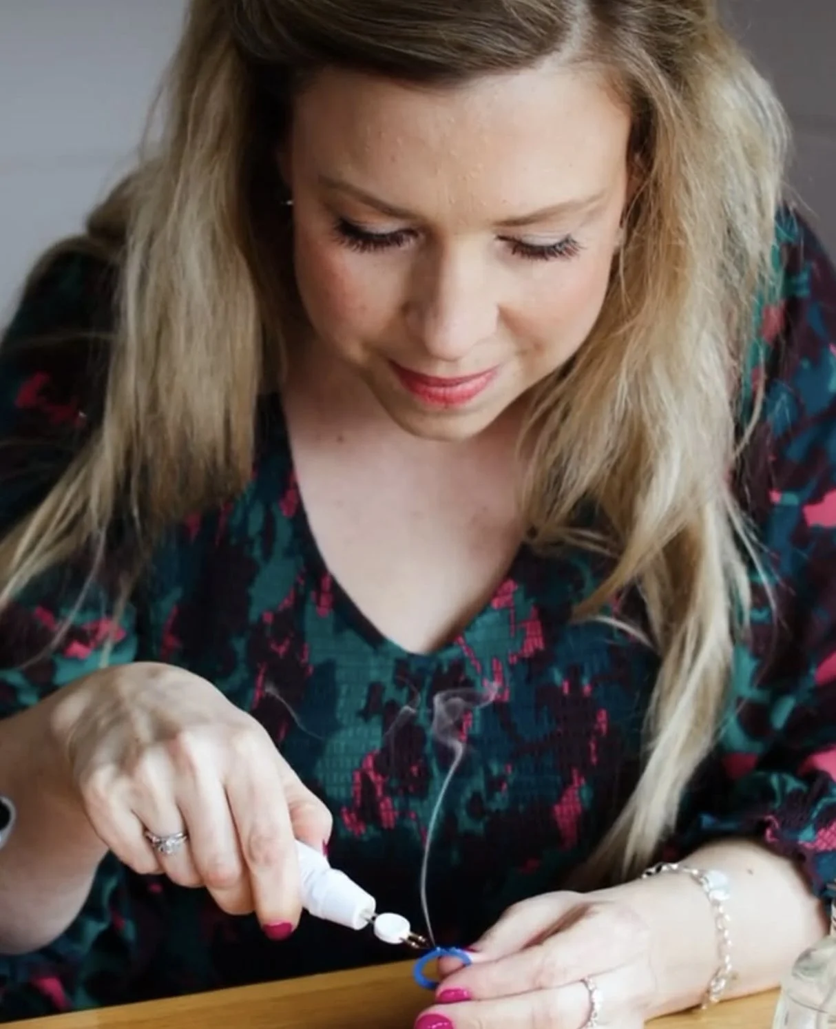 A woman with long blonde hair and a colorful patterned top is sitting at a wooden table, applying glue to a blue object.