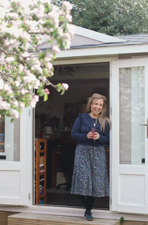 Anna Bagnall standing in her workshop, smiling and holding a cup, with pink and white flowers in the foreground.