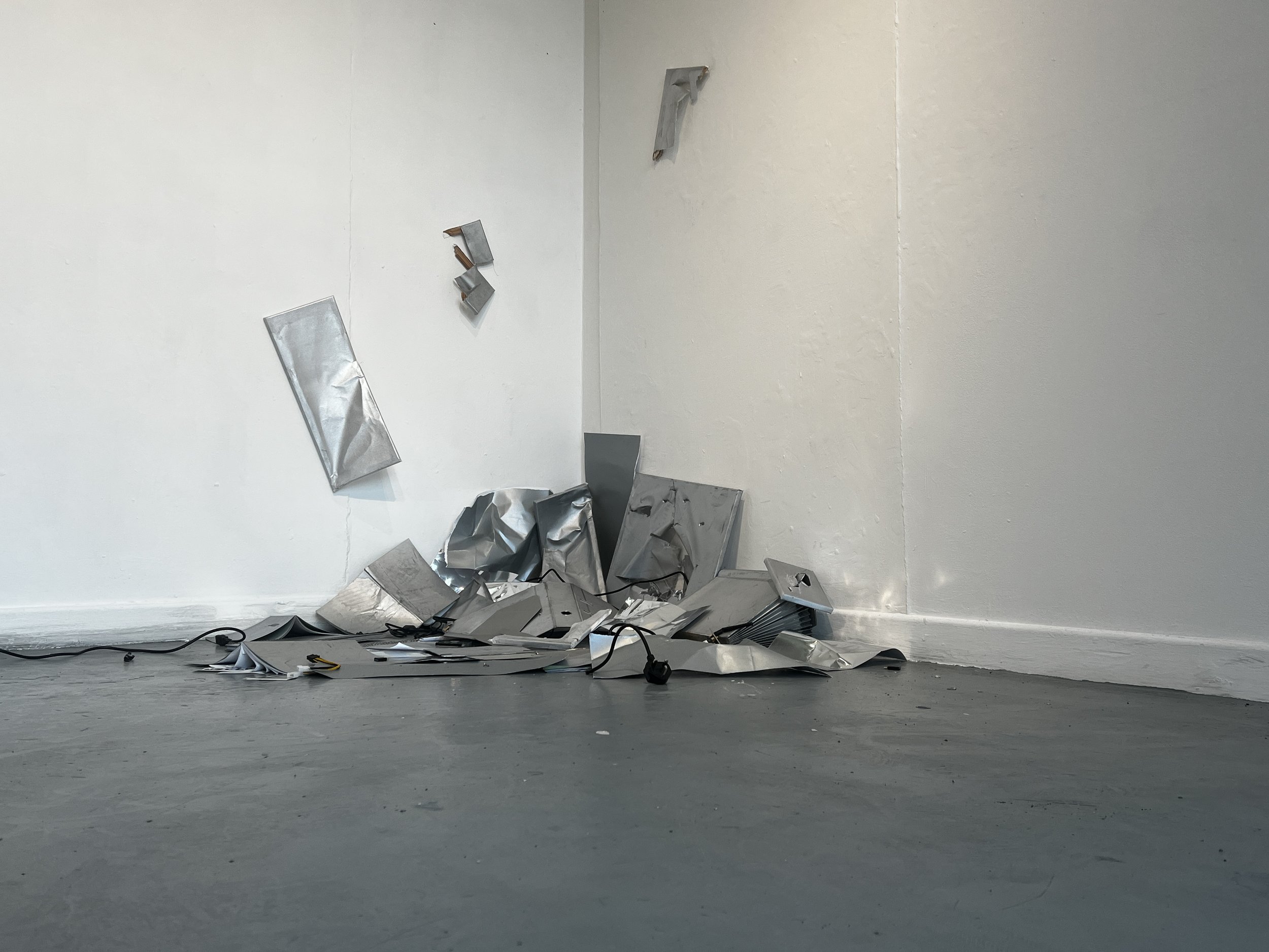 A room with white walls and a gray floor, scattered with crumpled and bent aluminum foil sheets, some hanging on the wall, and a black power cord on the floor.