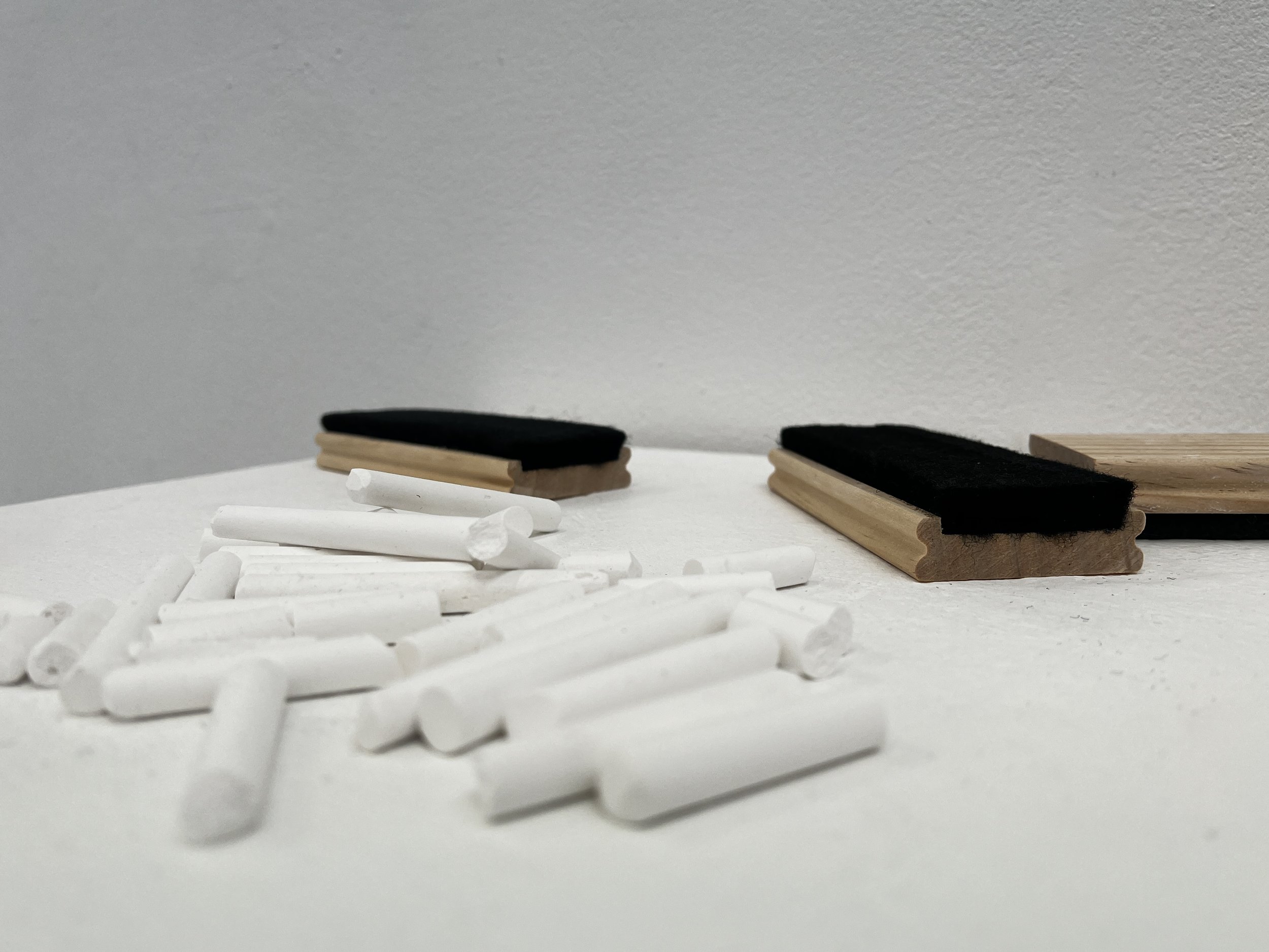 Close-up of a collection of white chalk sticks and wooden chalk holders on a white surface, with black felt-covered objects on top of the holders, against a plain light gray background.