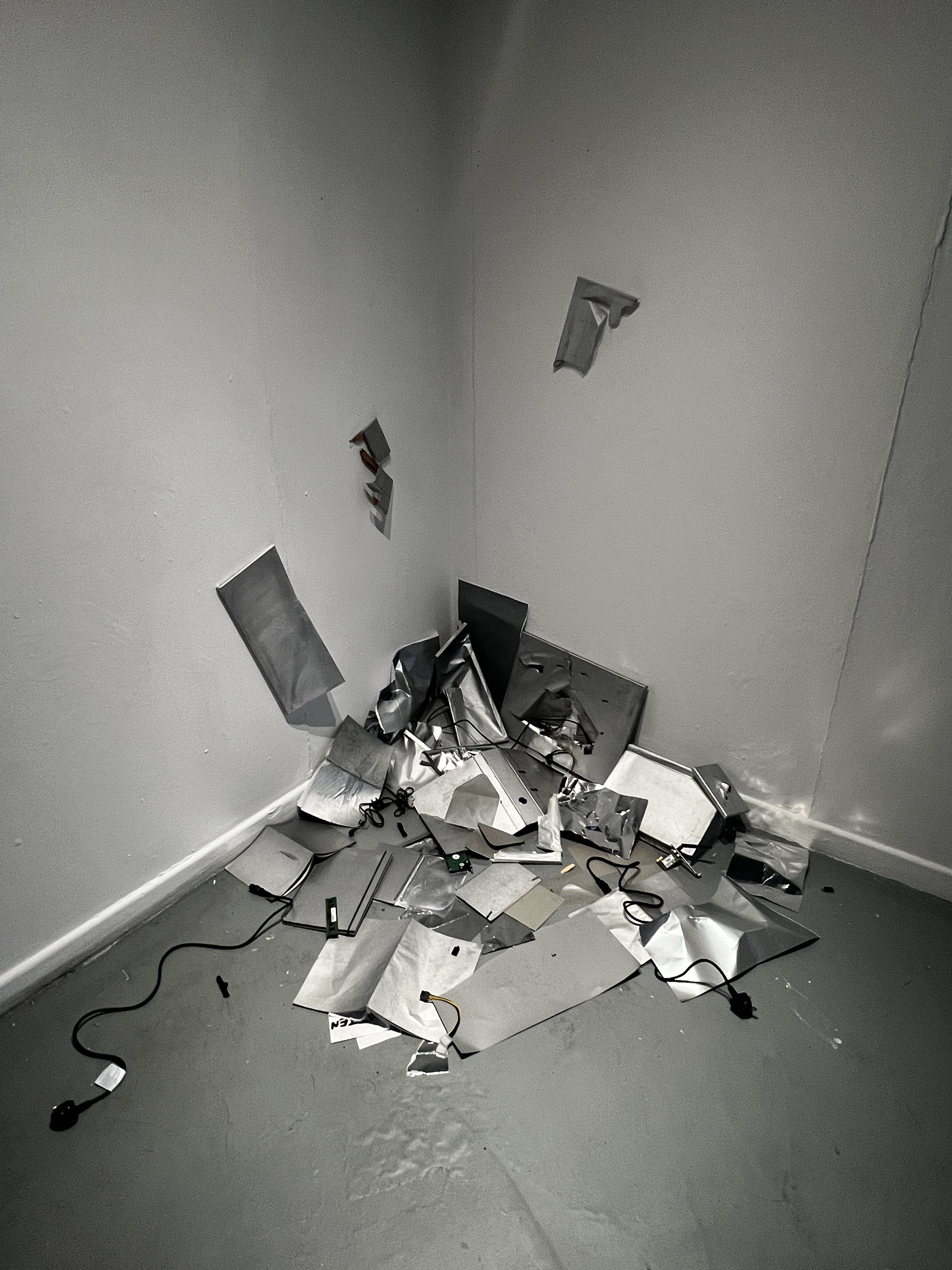 A corner of a room with a gray wall and floor, with broken pieces of silver metallic and white material, scattered on the floor and the wall, with some black electrical cords among the debris.