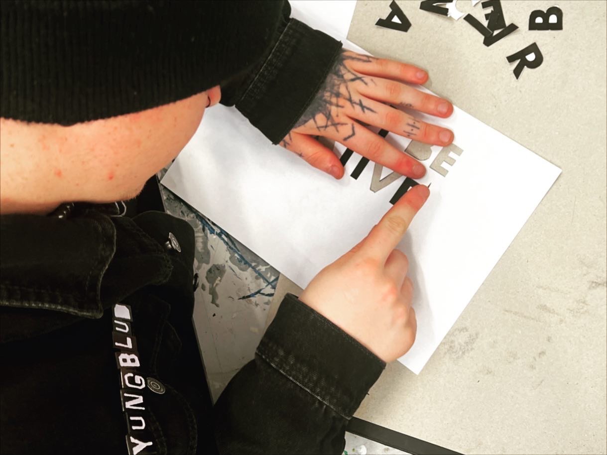 A person with tattoos on hands points at cut-out letters on a white sheet of paper. The person is wearing a black jacket and a black beanie, and has freckles on their face.