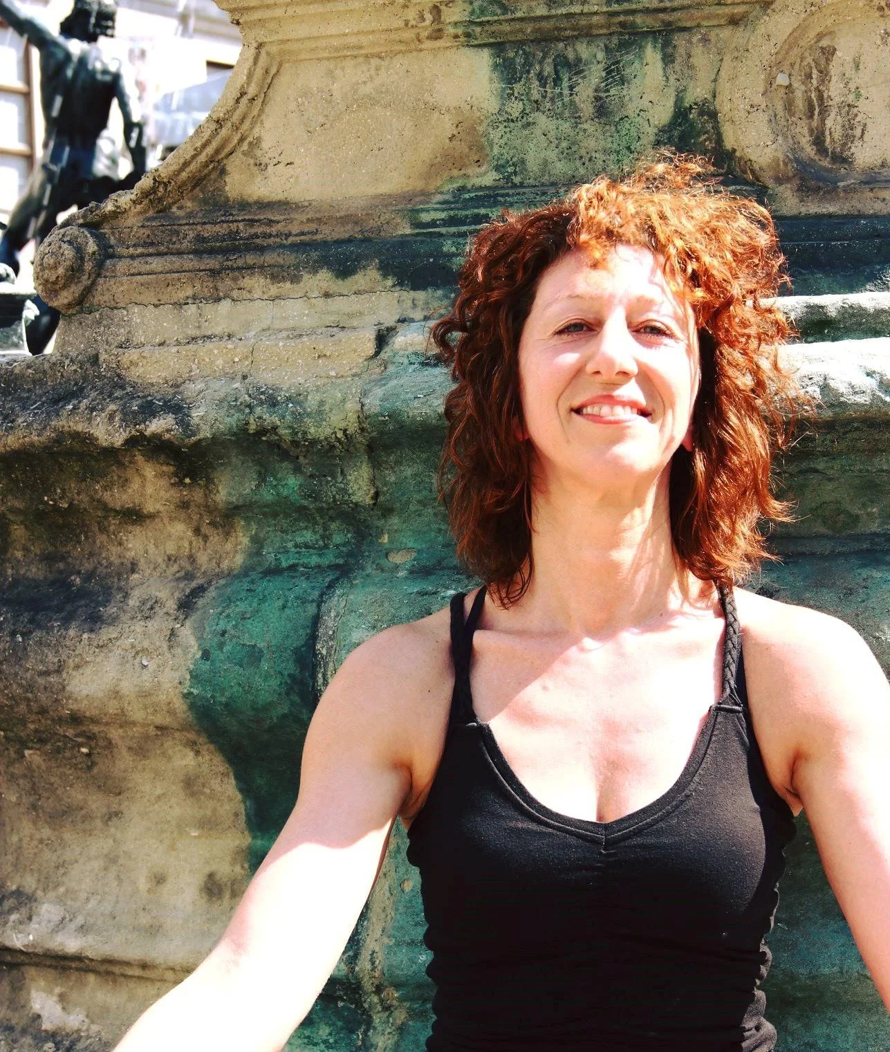 Eine Frau mit dunkelroten, lockigen Haaren trägt ein schwarzes Tanktop und lächelt, während sie vor einem steinernen Brunnen steht.