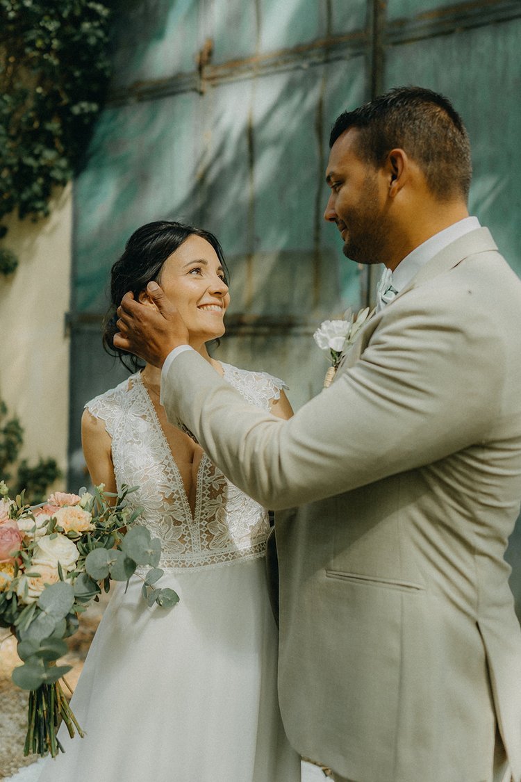 Un couple en tenue de mariage, l'homme touchant le visage de la femme, qui tient un bouquet de fleurs.