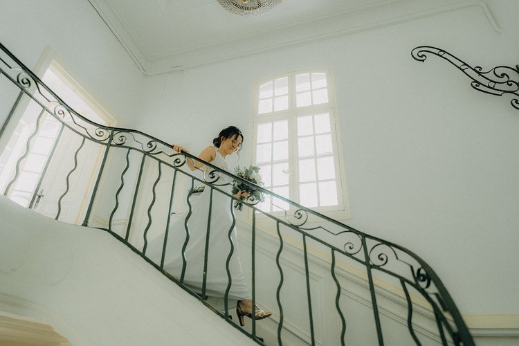 Femme en robe blanche descendant un escalier intérieur élégant, tenant un bouquet de fleurs.