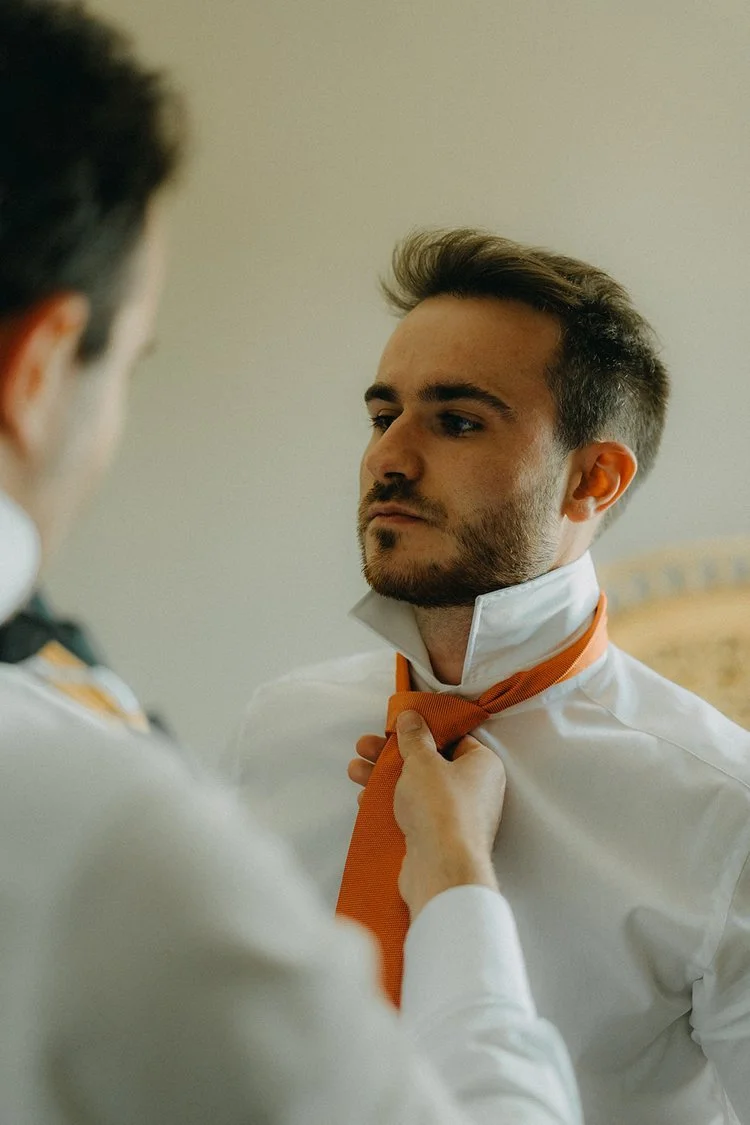 Un homme ajustant une cravate orange sur une chemise blanche devant un miroir.