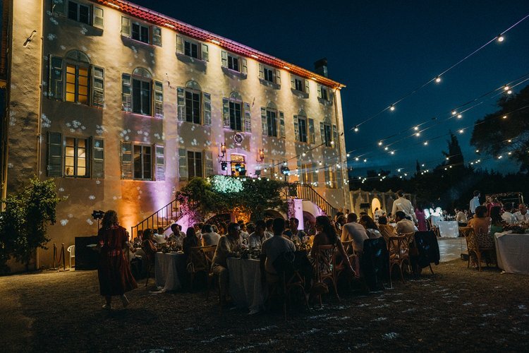 Réception en plein air avec des invités assis à des tables élégantes, devant un bâtiment illuminé, sous des guirlandes lumineuses.