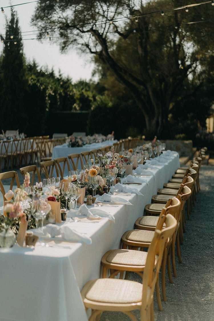 Tables de réception en plein air décorées de fleurs et de verres, avec des chaises en bois, sous un arbre et des guirlandes lumineuses.