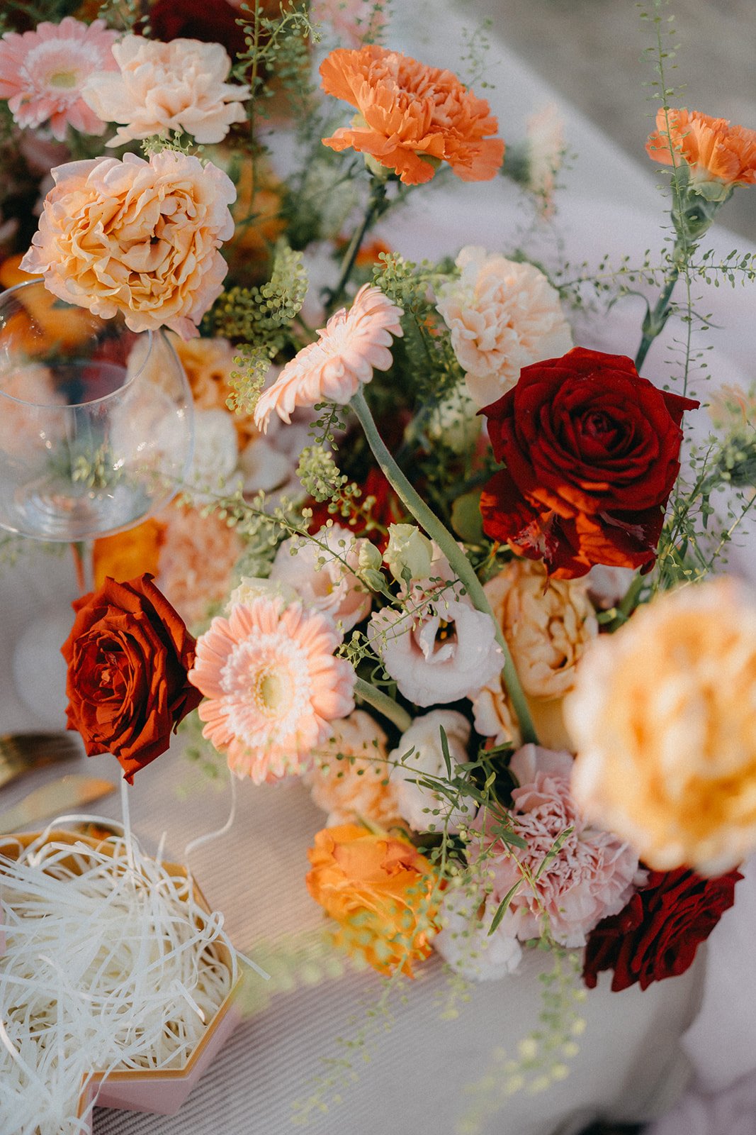 Arrangement de fleurs avec roses rouges, œillets orange et roses, sur une table avec verres à vin.