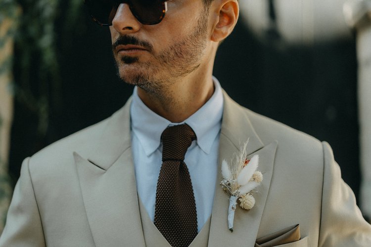 Un homme portant un costume beige, une chemise blanche et une cravate marron, avec des lunettes de soleil. Une boutonnière de fleurs séchées est attachée à sa veste.