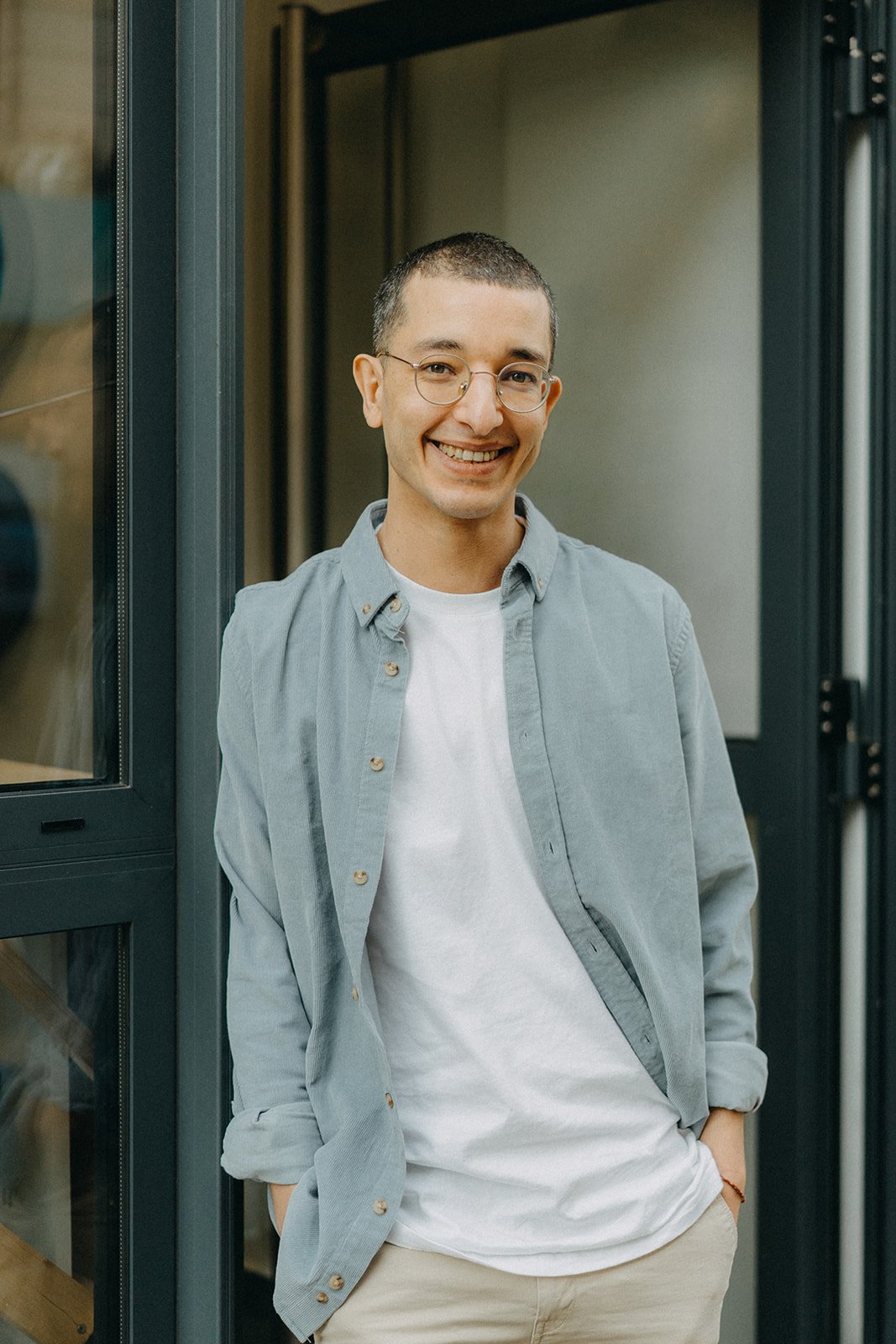 Un homme souriant portant des lunettes, une chemise ouverte sur un t-shirt blanc, debout devant une porte, les mains dans les poches.