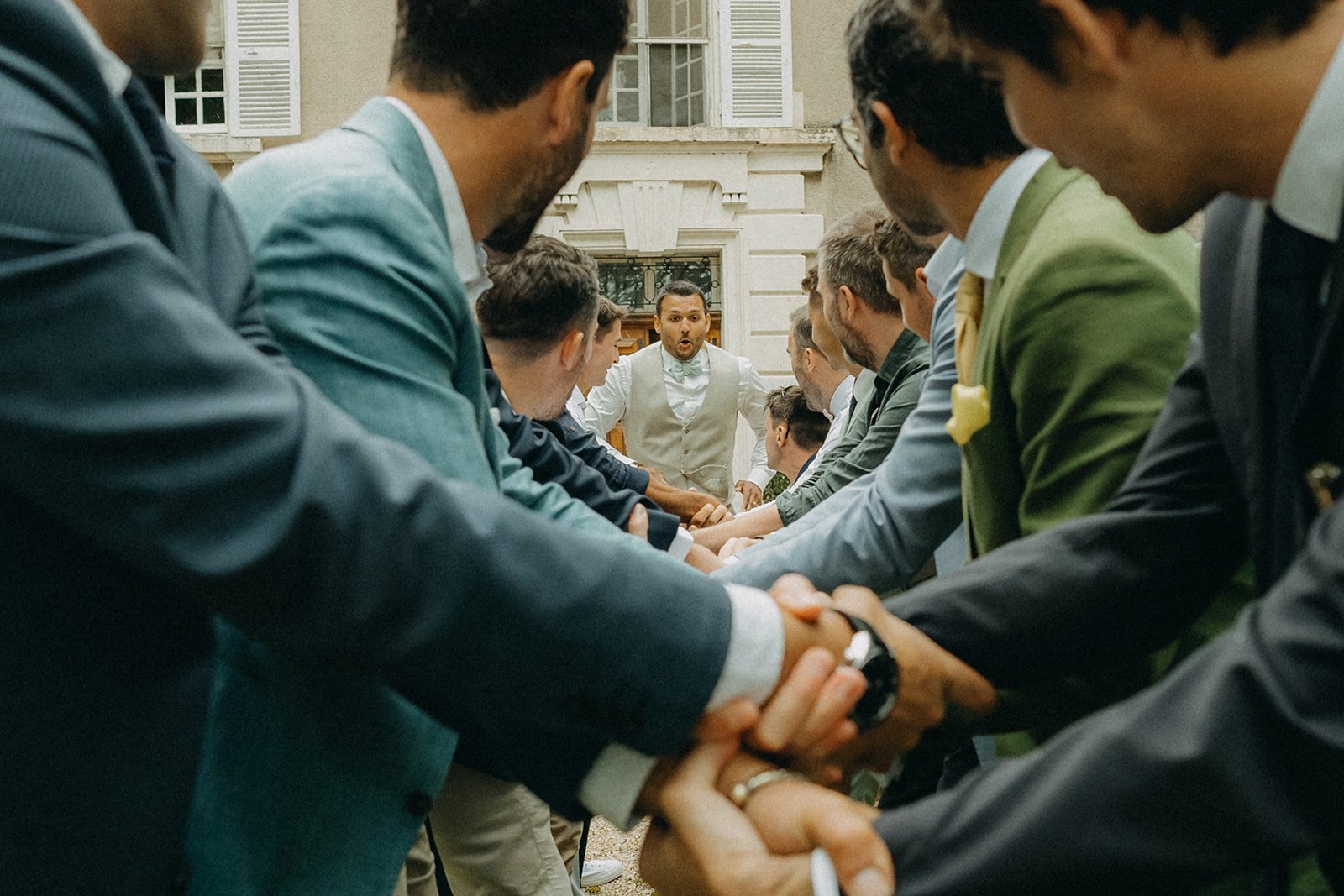 Un groupe d'hommes en costume se tient par les mains, formant un tunnel, avec un homme au centre faisant une expression surprise. La scène semble se dérouler lors d'une célébration à l'extérieur.