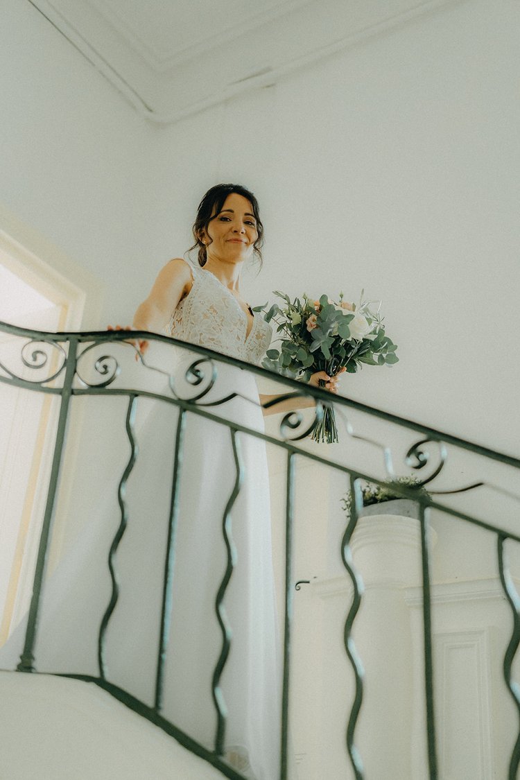 Une femme en robe de mariée tenant un bouquet, debout dans un escalier.