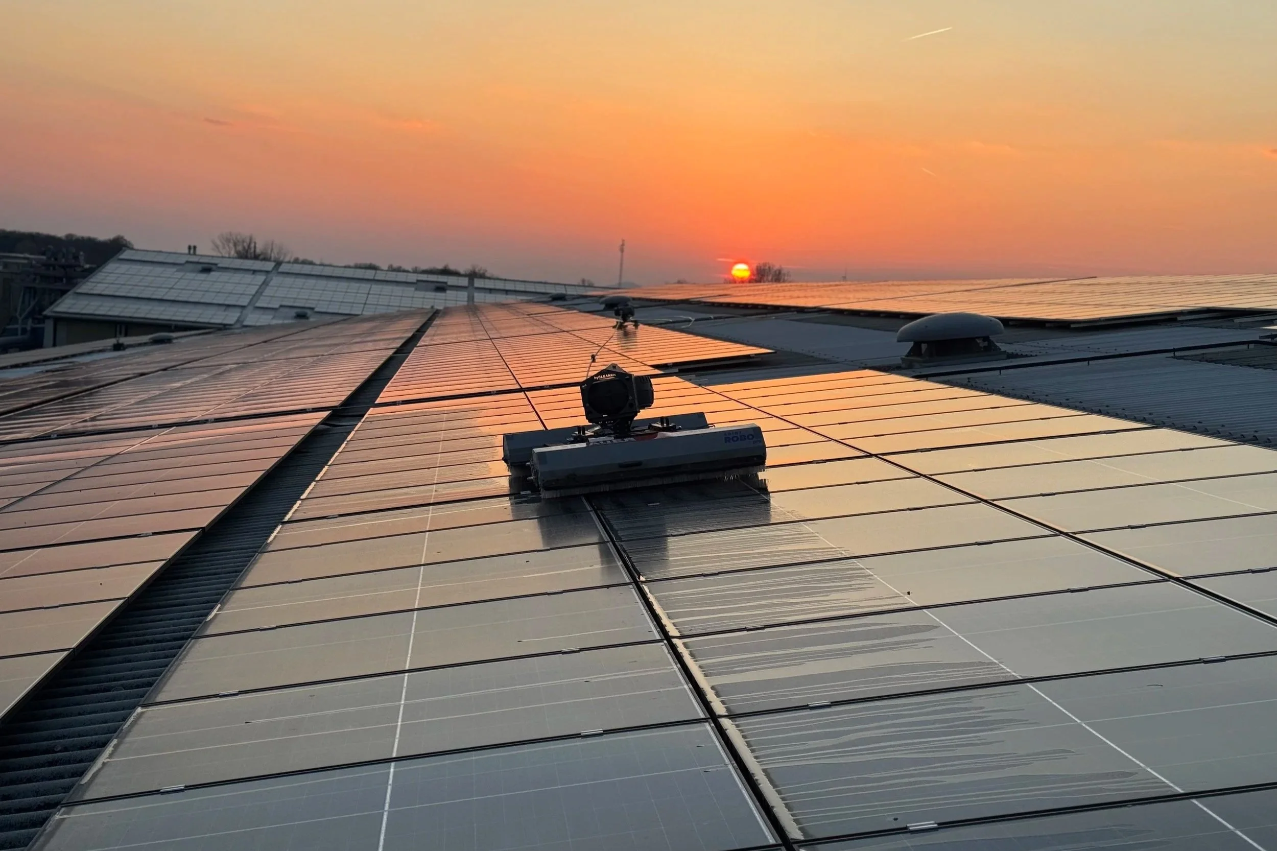 Ein Solarreinigungsroboter reinigt eine Photovoltaikanlage auf einem Dach, während im Hintergrund ein stimmungsvoller Sonnenuntergang zu sehen ist. Die moderne Reinigungstechnik sorgt für eine effiziente und schonende Reinigung der Solarmodule.
