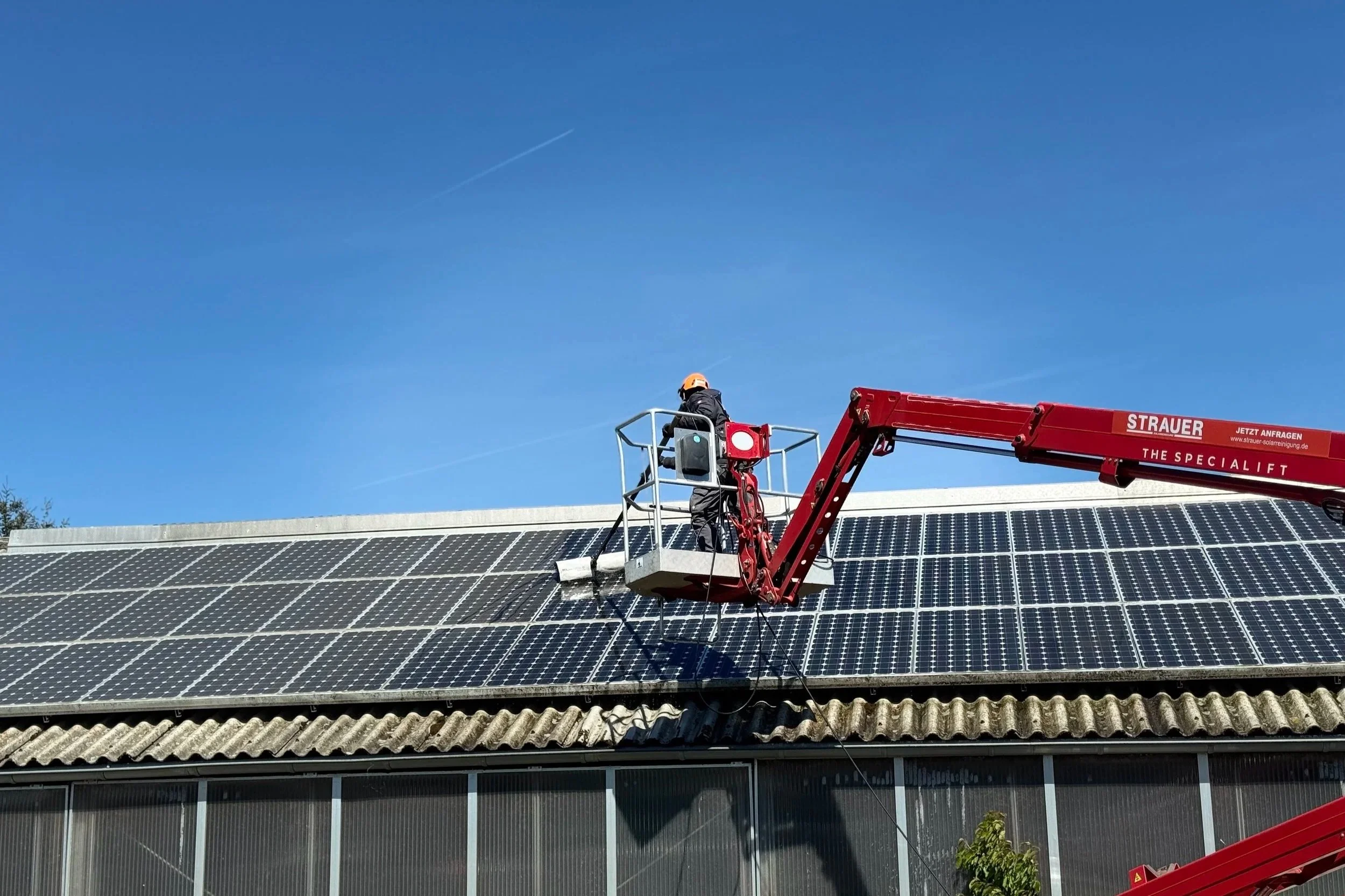 Ein Solarreiniger säubert eine Solaranlage auf einem Dach aus dem Steigerkorb einer Arbeitsbühne mit einer rotierenden Reinigungsbürste. Die professionelle Solarreinigung entfernt Verschmutzungen schonend und trägt zur langfristigen Leistungsfähigkei