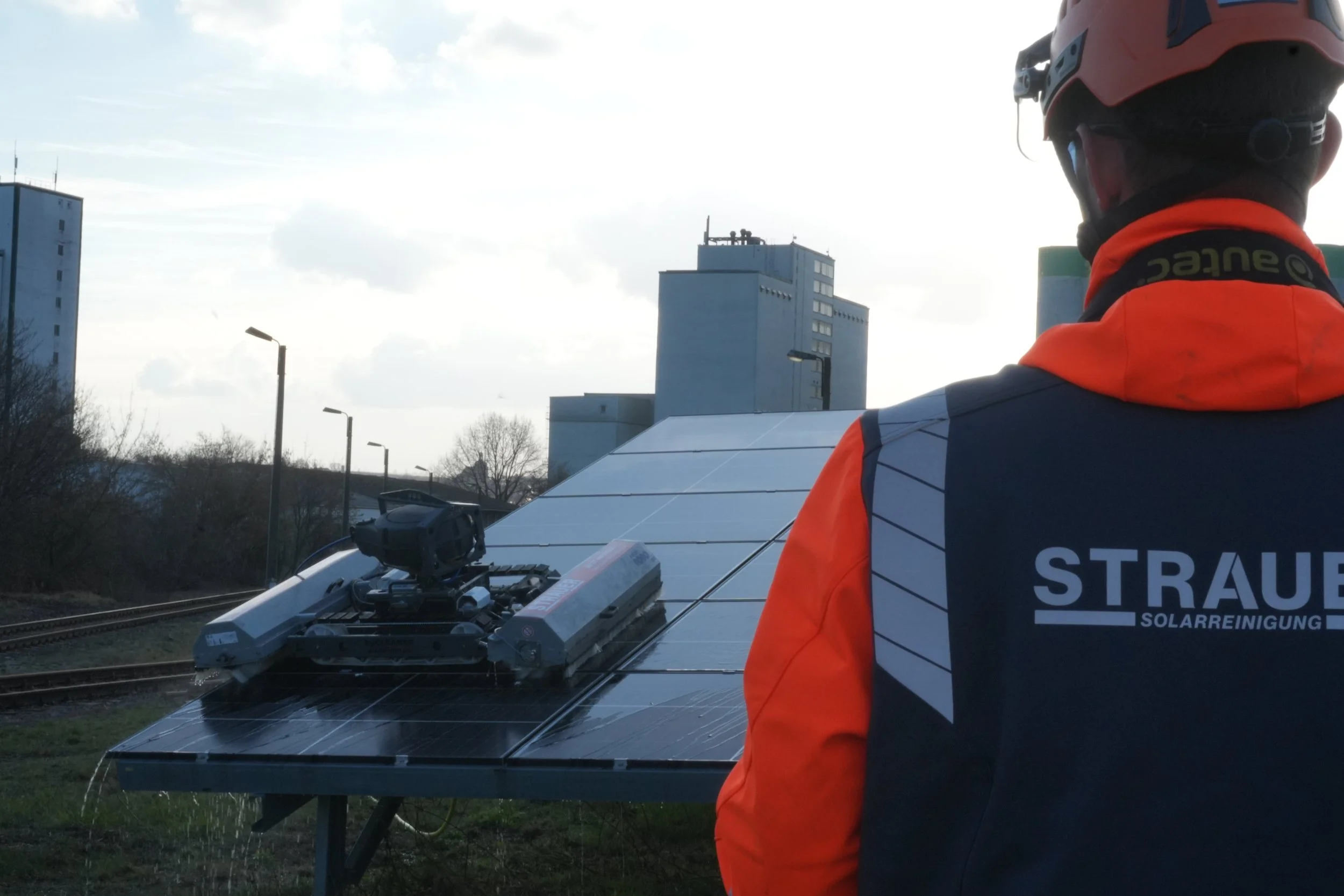 Ein STRAUER-Solarreiniger steht vor einer Freiflächenanlage, während im Hintergrund ein Solarreinigungsroboter die Solarmodule effizient reinigt. Das Zusammenspiel aus erfahrenem Fachpersonal und moderner Reinigungstechnik ermöglicht eine professione