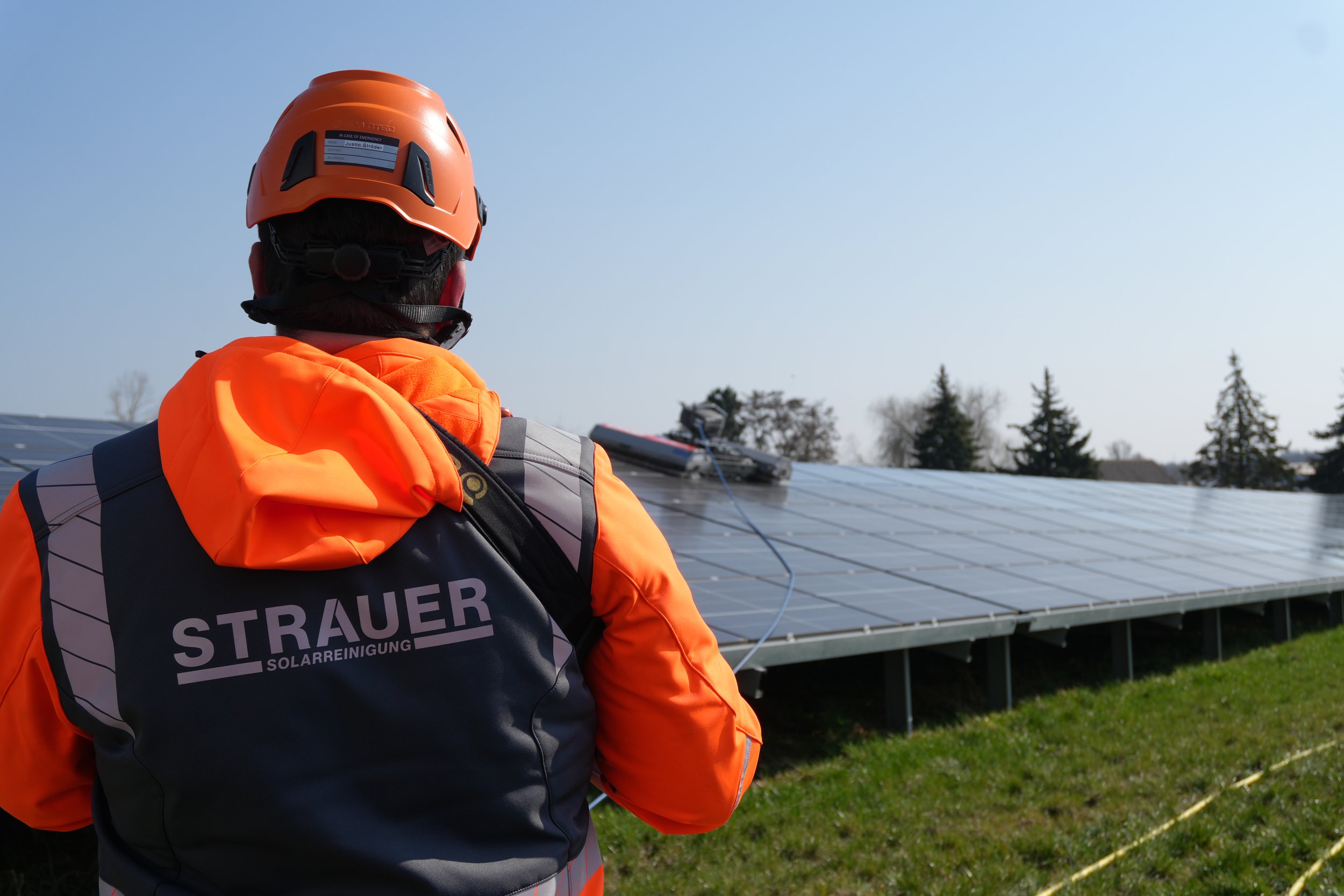 Ein_Mann_steht_mit_Strauer-Arbeitsbekleidung_vor_einem_Solarfeld_und_im_Hintergrund_ist_ein_Reinigungsroboter.png