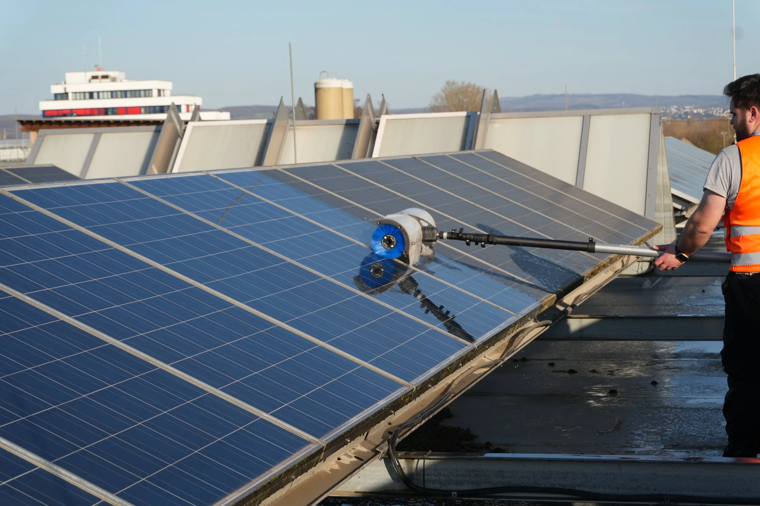Ein Techniker reinigt Solarpaneele auf einem Dach mit einer Hochdruckreinigungsmaschine an einem sonnigen Tag.