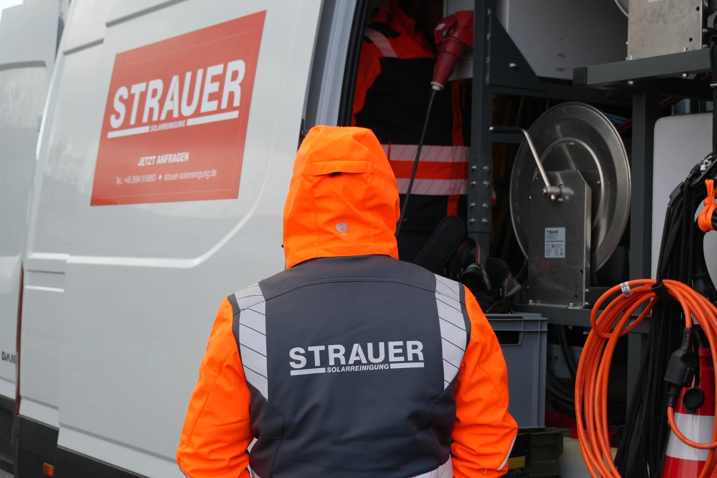 Ein Techniker der Firma 'Strauer Solarreinigung' trägt eine orange und schwarze Jacke mit dem Logo auf dem Rücken, steht vor einem Servicefahrzeug mit geöffnetem Fach, in dem Reinigungsgeräte und Kabel für die Solaranlagenreinigung aufbewahrt werden.