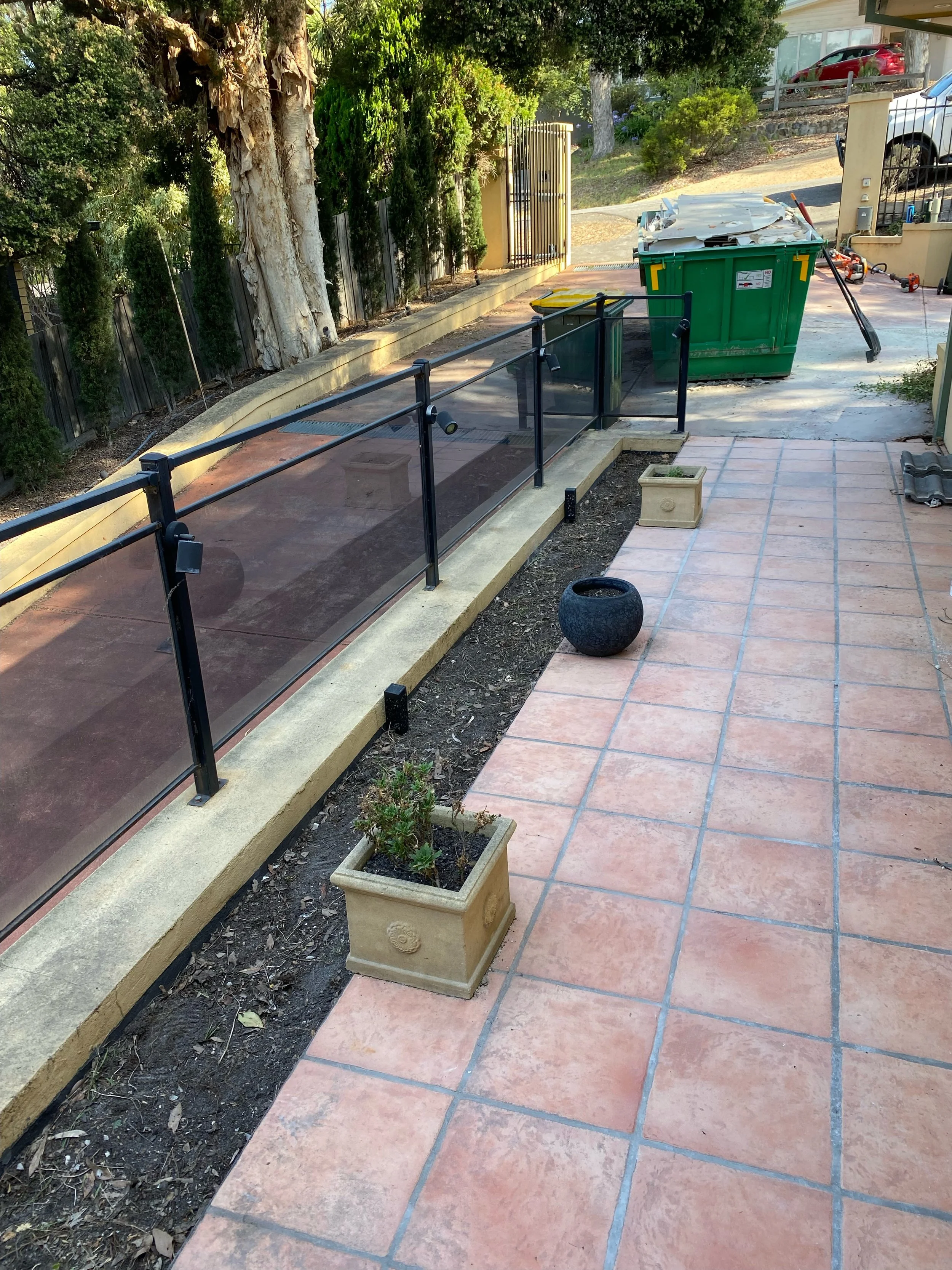 Outdoor patio with terracotta tiles, black metal railing, small potted plants, and a green dumpster in the background.