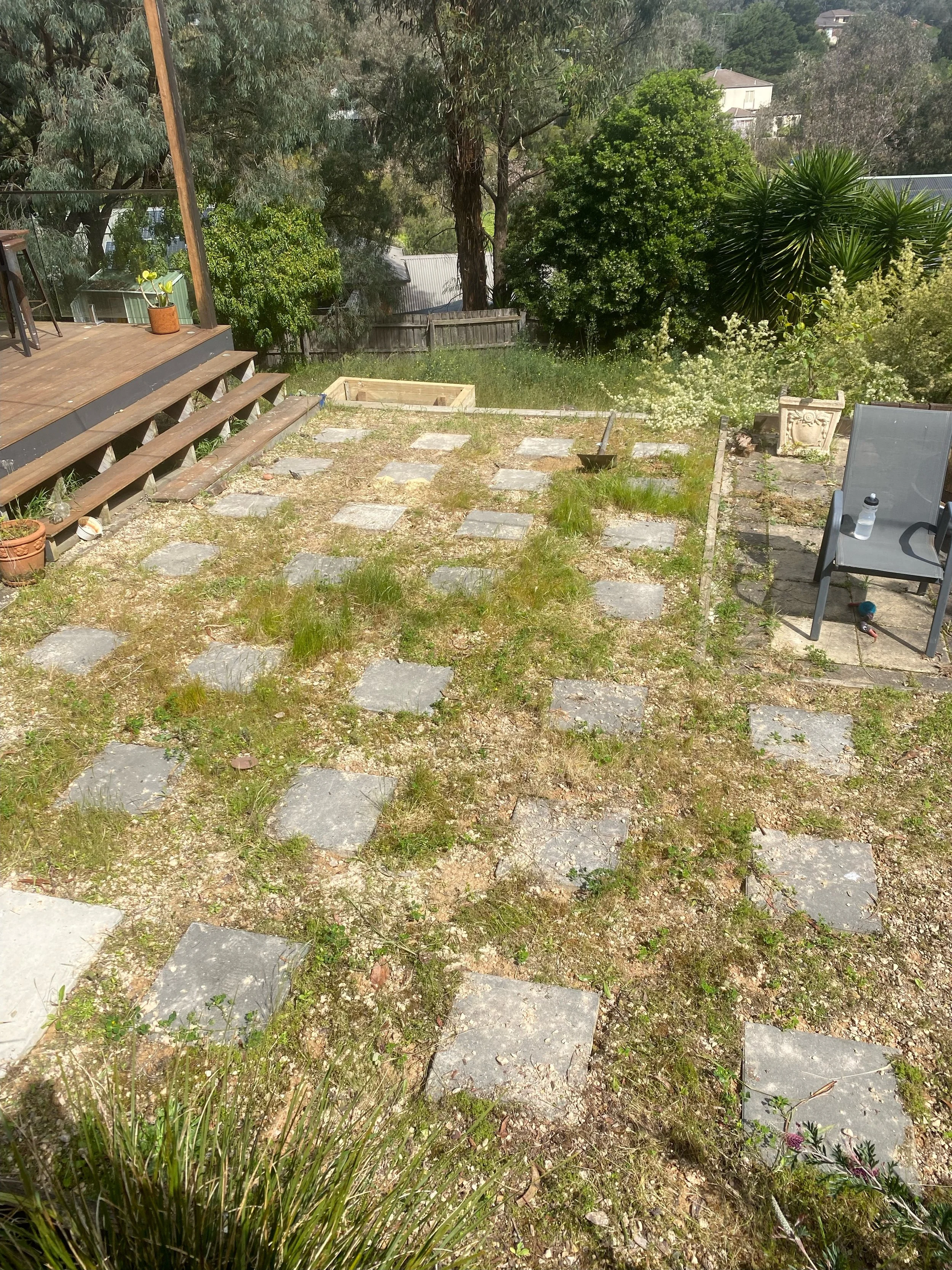Backyard garden with square stone pavers on a dirt and grass surface, surrounded by trees and shrubs. Wooden deck with stairs is visible, along with an outdoor chair and a potted plant.