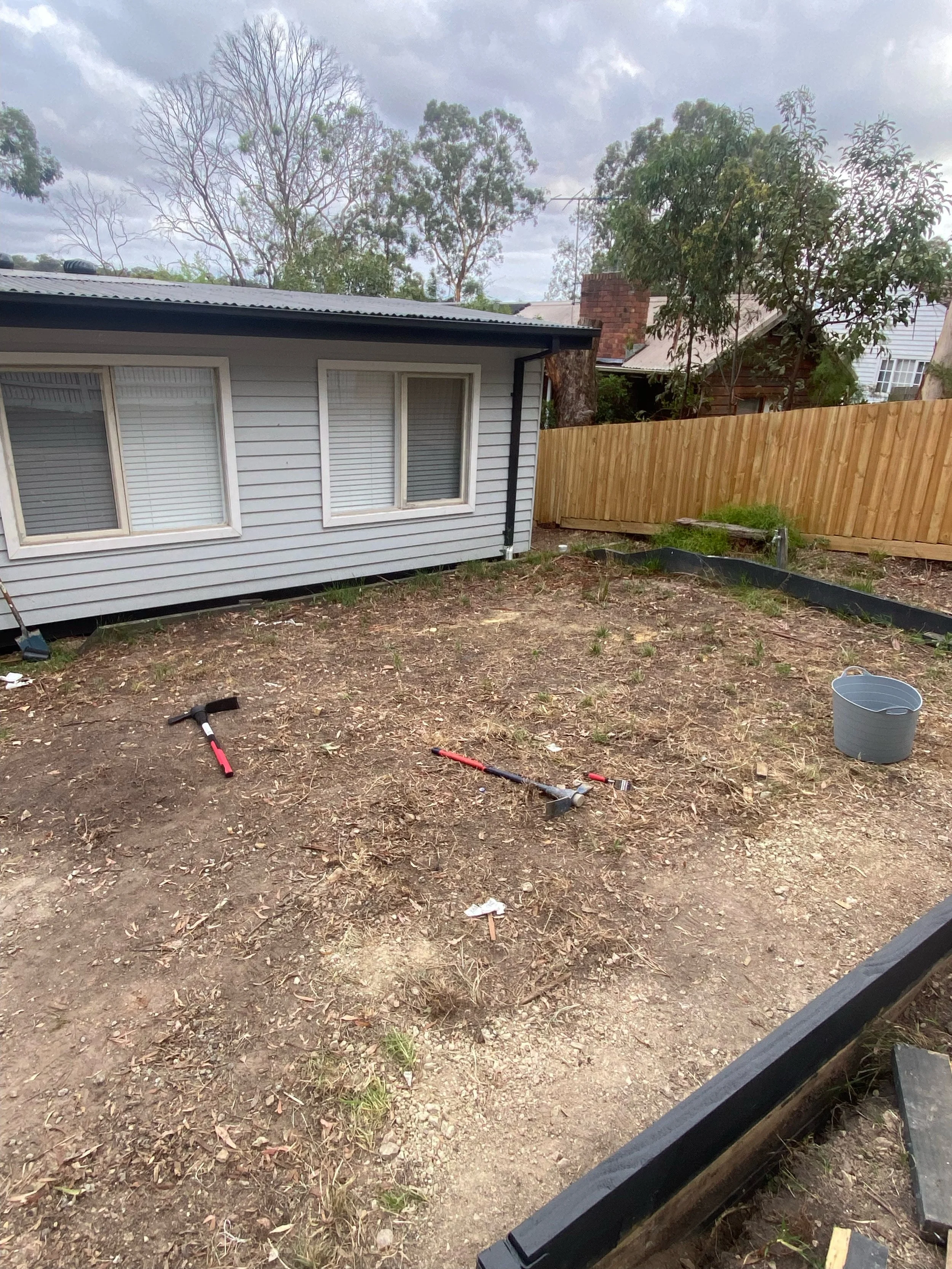 Backyard with a wooden fence, small house, bare soil, gardening tools, and a bucket.