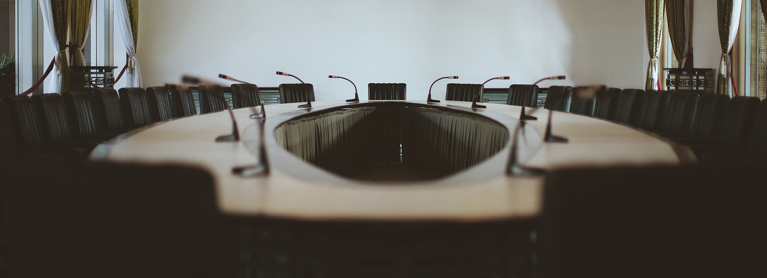 Large empty conference table with numerous microphones and chairs, set in a room with draped windows and decorative elements.