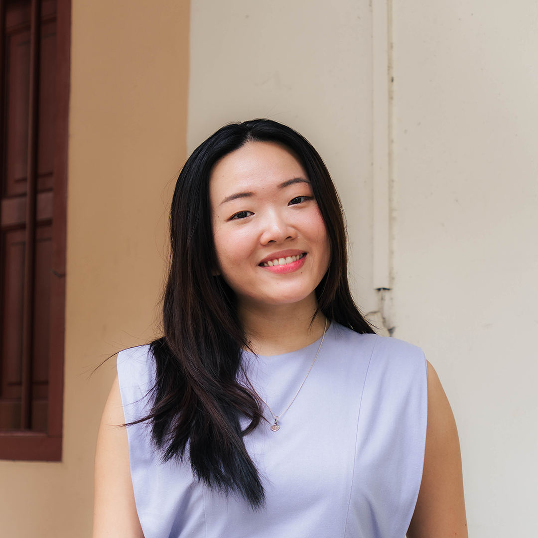 Smiling woman in a light blue sleeveless top with a pendant necklace, standing against a neutral background.