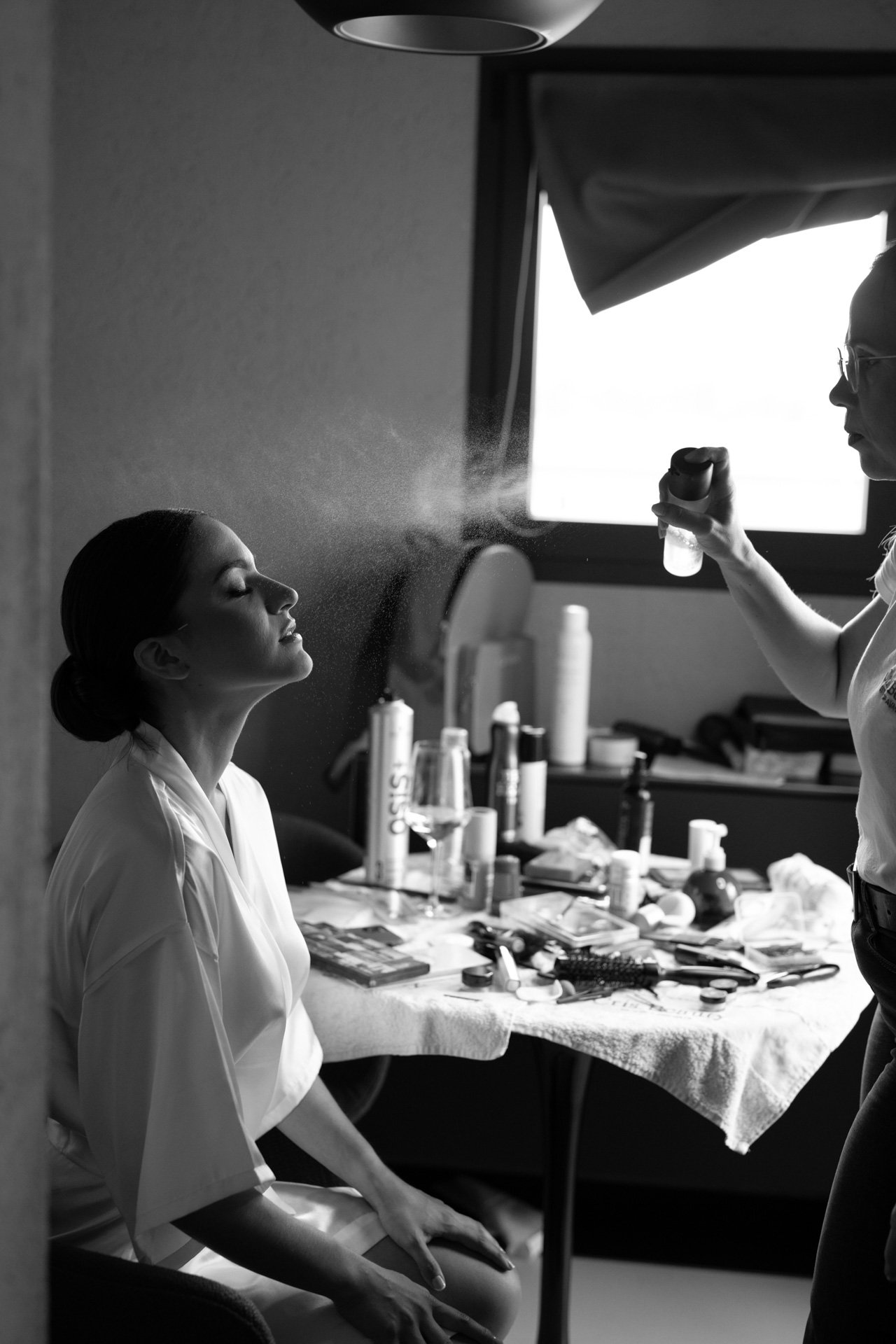 Una persona seduta su una sedia mentre un'altra persona applica spray per il viso. Sul tavolo ci sono vari prodotti cosmetici e accessori per il trucco.